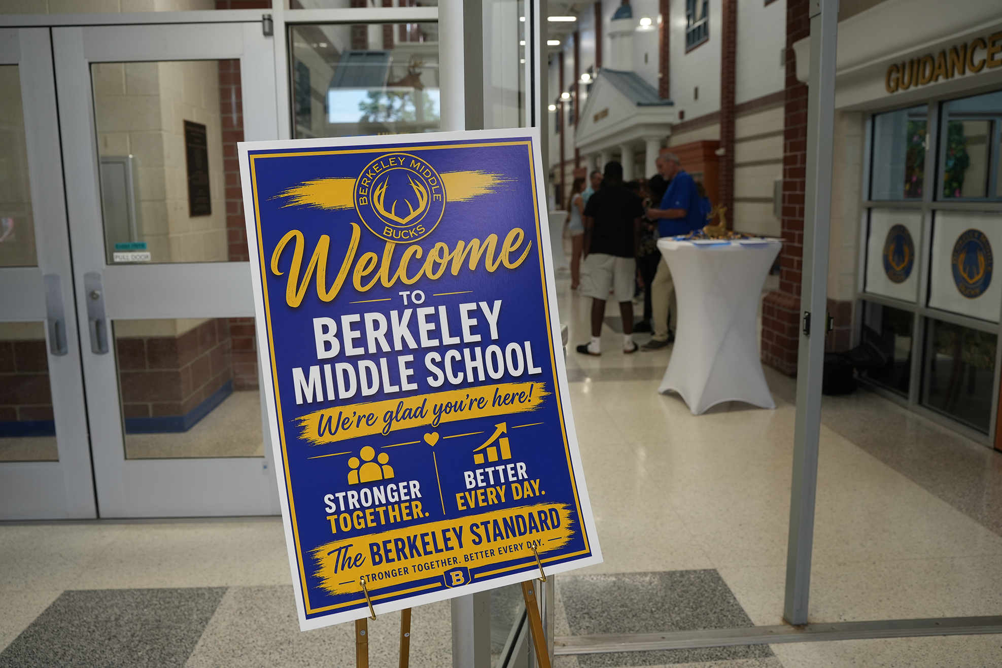 A blue and yellow sign reading "Welcome to Berkeley Middle School" in a school hallway with people and a pillar.