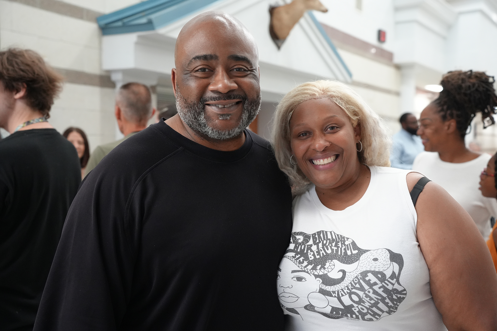 A man and a woman smiling, standing together with others in a hallway. The man wears a black shirt; the woman, a white shirt.