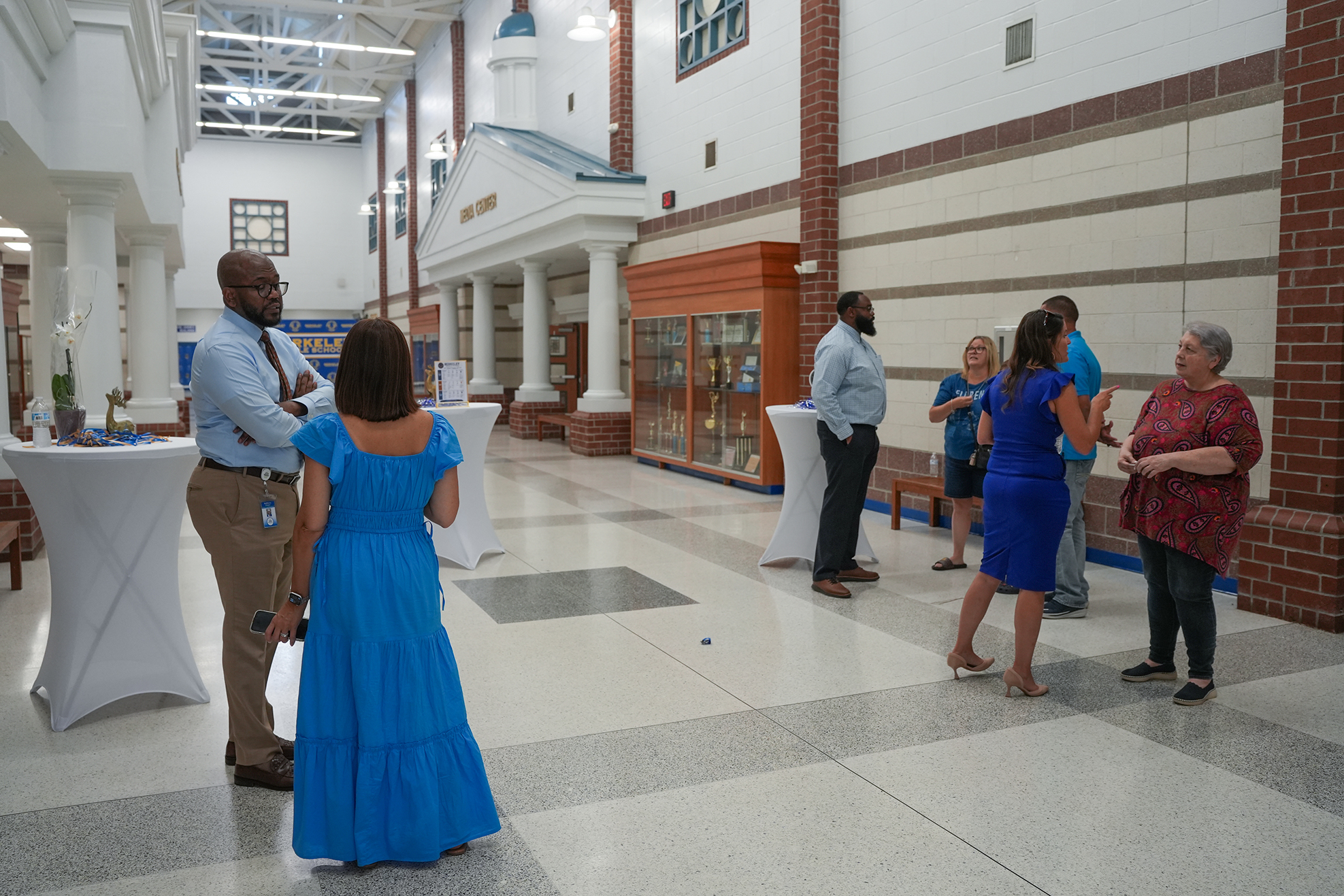 Nine adults in a hallway with white and brick walls, engaged in conversation. White tables are positioned at the left.