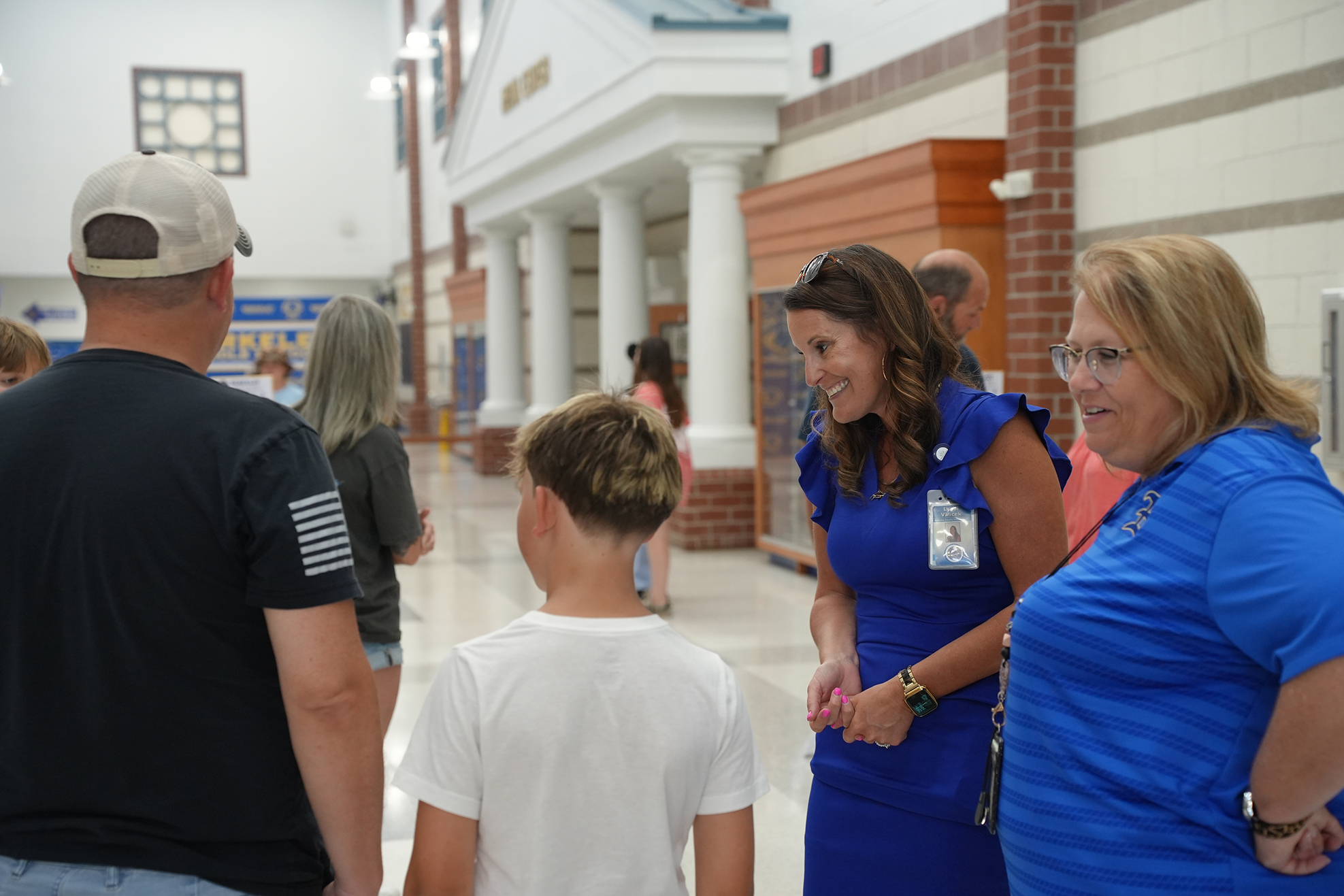 Several adults and children are standing in a hallway. A woman with glasses is speaking to a boy.