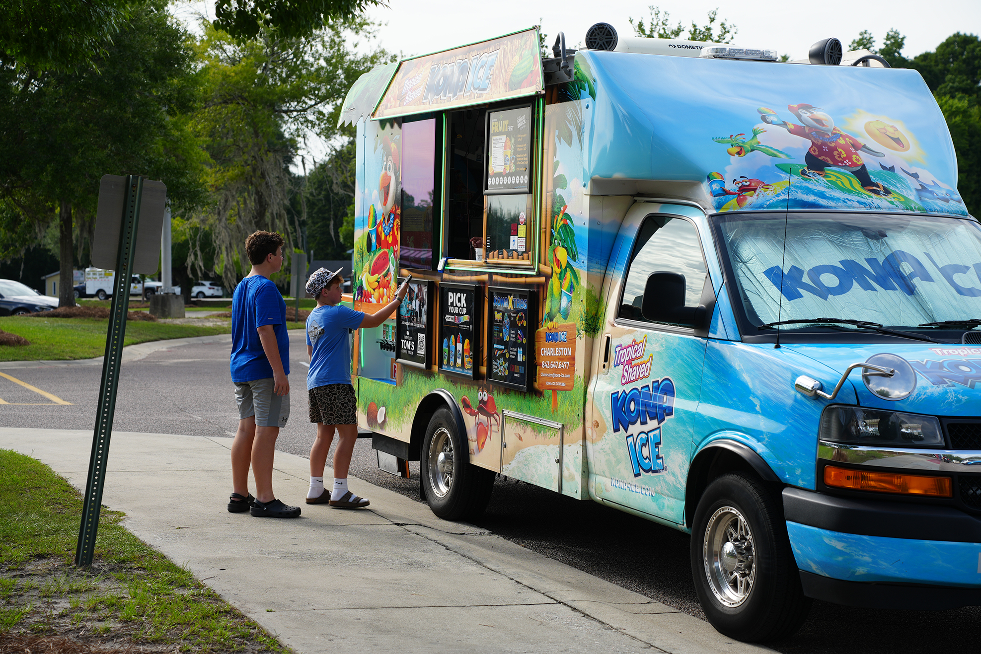 Two individuals, one with a hat, are at a colorful ice cream truck on a sidewalk.
