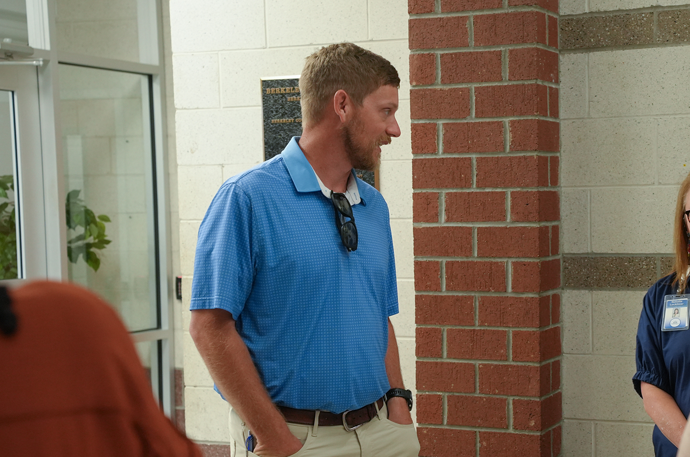 A man in a blue shirt stands by a brick wall, looking to his right. Another person is partially visible.