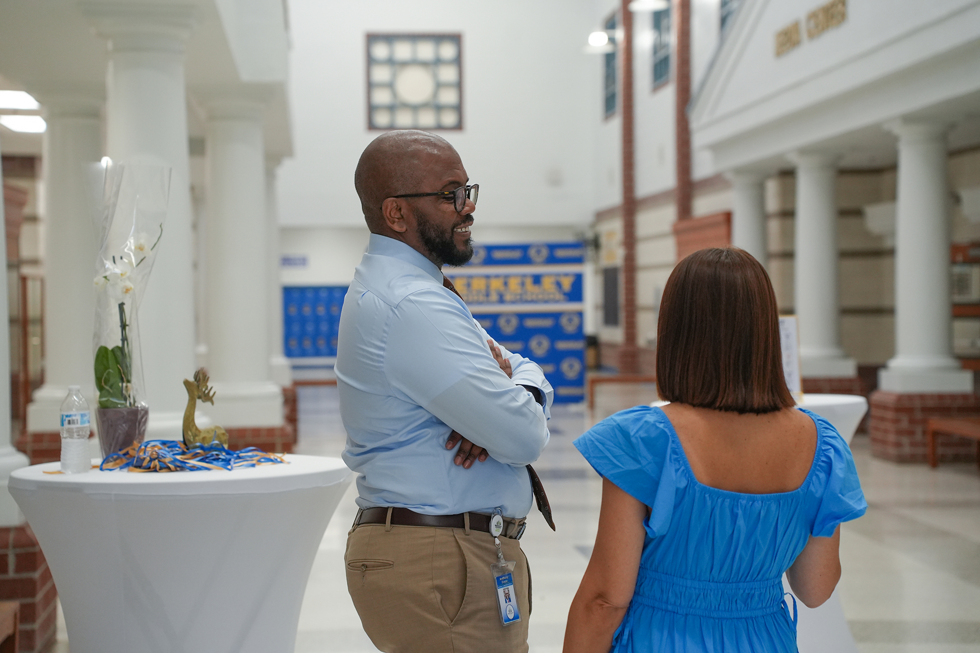 A man in a light blue shirt and khaki pants stands with his arms crossed. A woman in a blue dress stands opposite him.