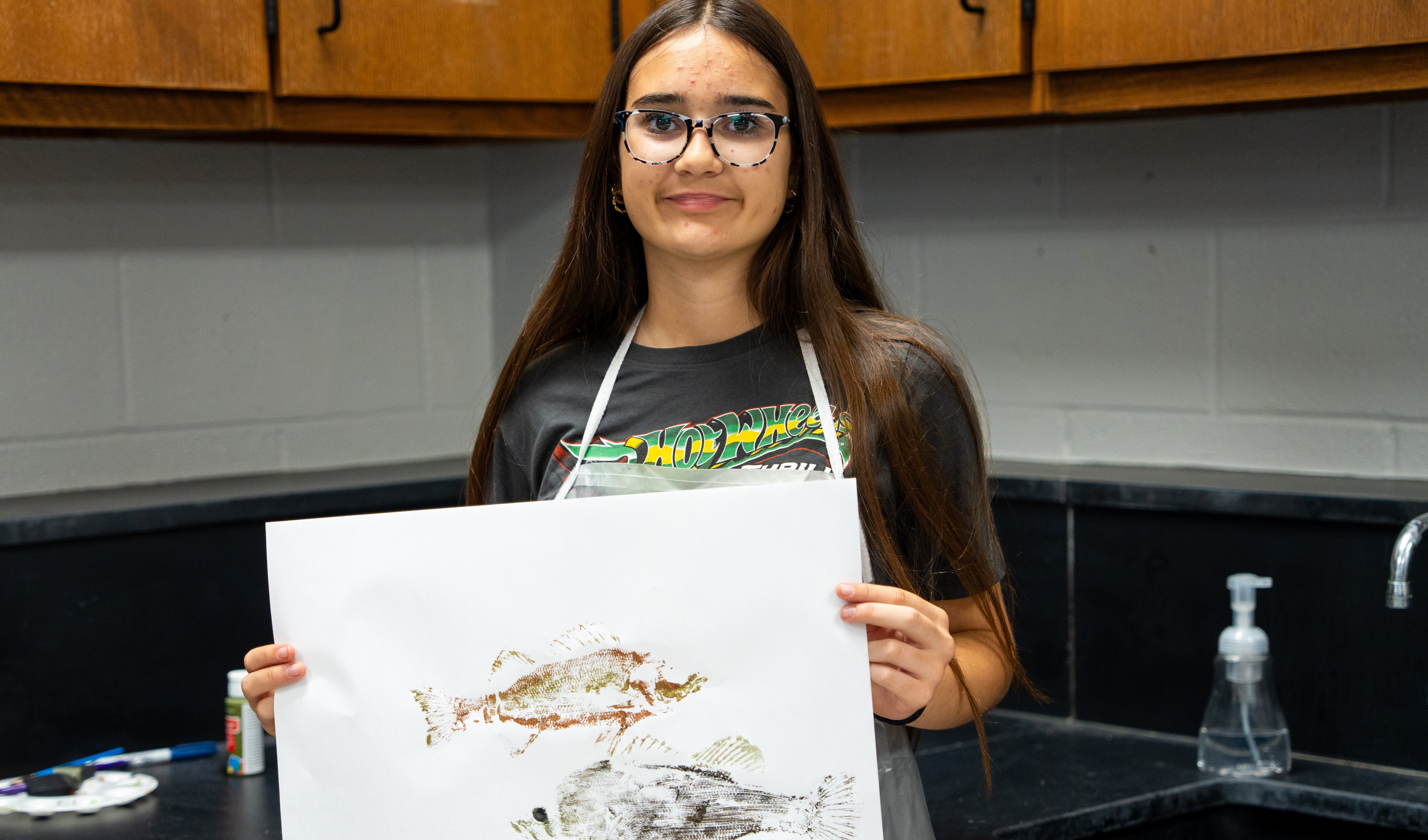 Person wearing glasses and an apron holds up a white paper with a fish drawing.