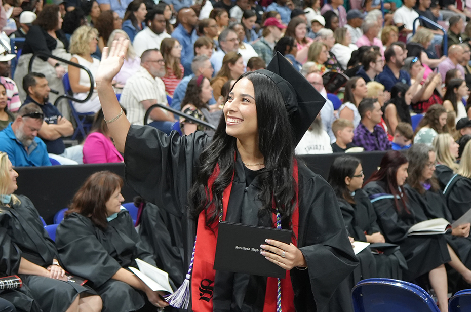 A smiling graduate in a black gown waves at the camera. She holds a diploma and stands among seated graduates and an audience.