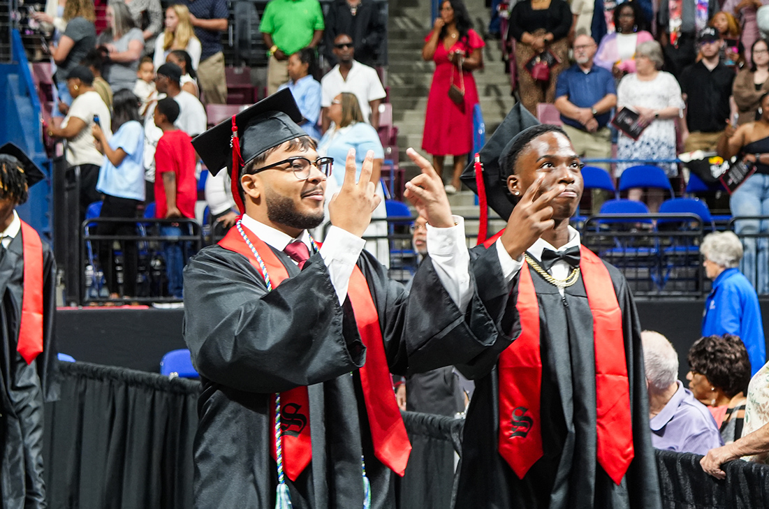 Two men in academic regalia celebrate on a stage with a large crowd of spectators in the background.