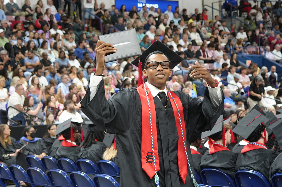 A man in a black gown with a red sash stands in a crowd of people, holding a diploma.
