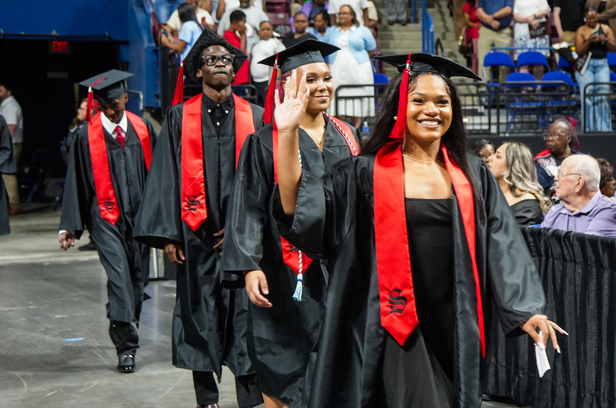 Four people in black gowns and hats walk in a procession. One woman smiles and waves. Spectators sit in stadium seating.