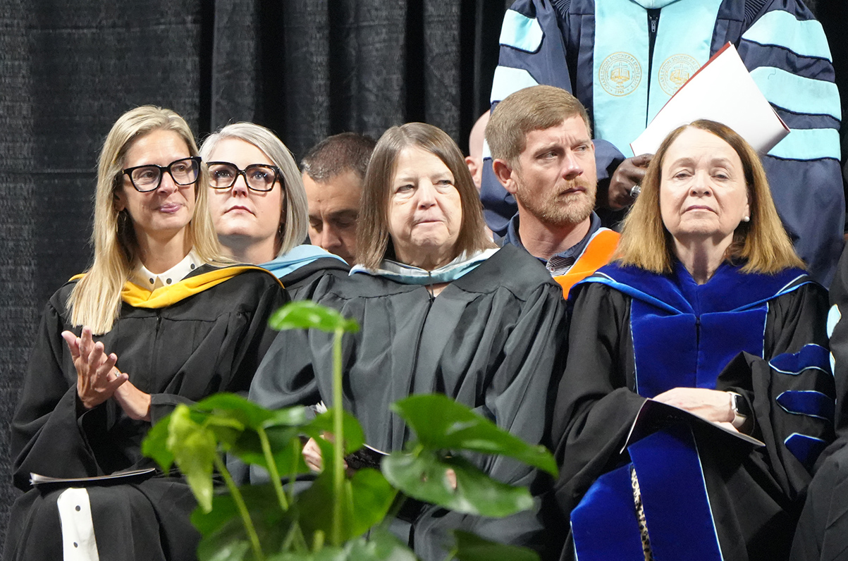 People in graduation robes sit at a ceremony. A plant is in the foreground.