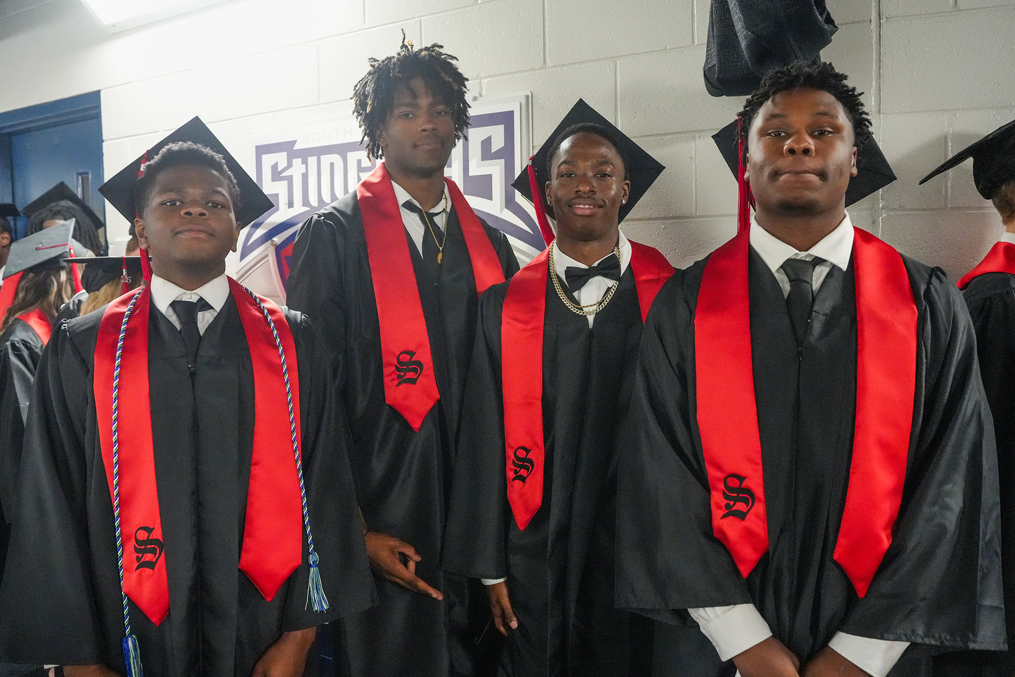 Five men in black graduation gowns and red stoles stand together, smiling for a photo in a hallway.