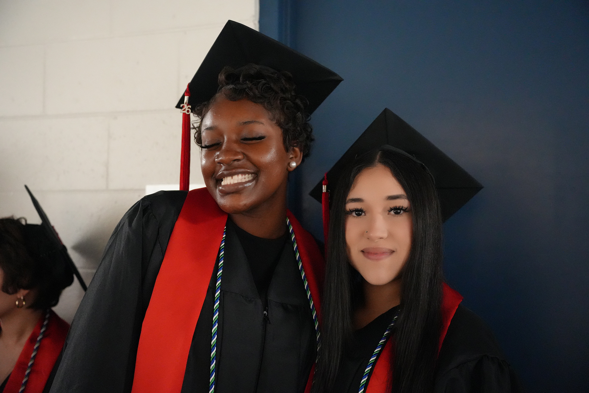Two young women in graduation robes and caps pose for a photo, one smiling. They wear sashes over black gowns.