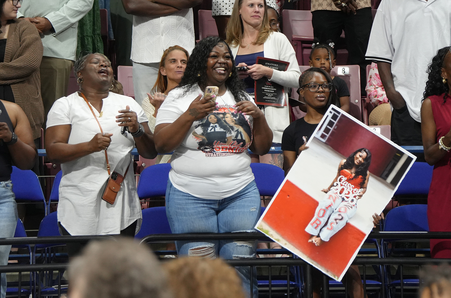 Spectators stand in a stadium, one woman holds a sign with a photo, another holds a phone, and others look on.