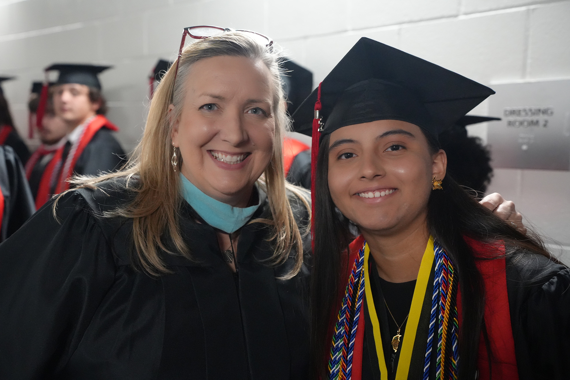 Two women, one in a graduation gown and cap, the other in a stole, stand smiling. Behind them, blurred images of others in graduation attire.