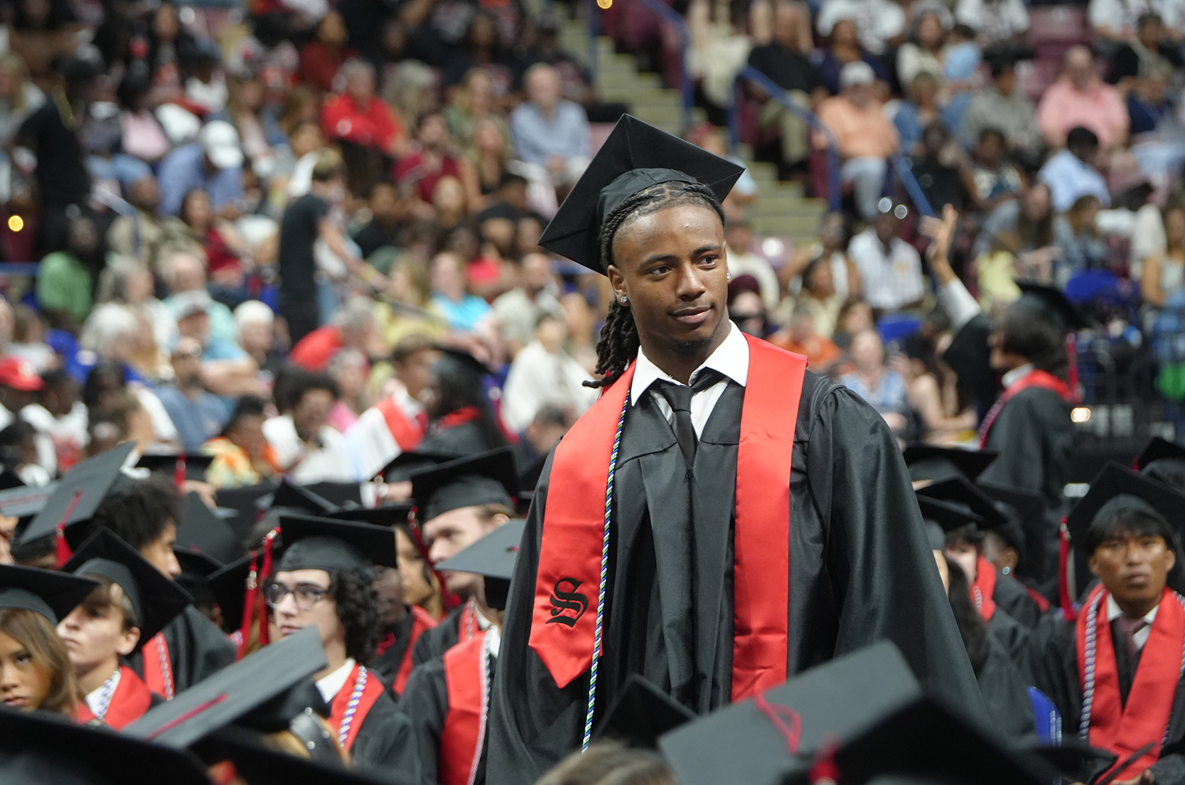 A person wearing a graduation gown and hat stands in a crowded auditorium. Many people are seated behind them.