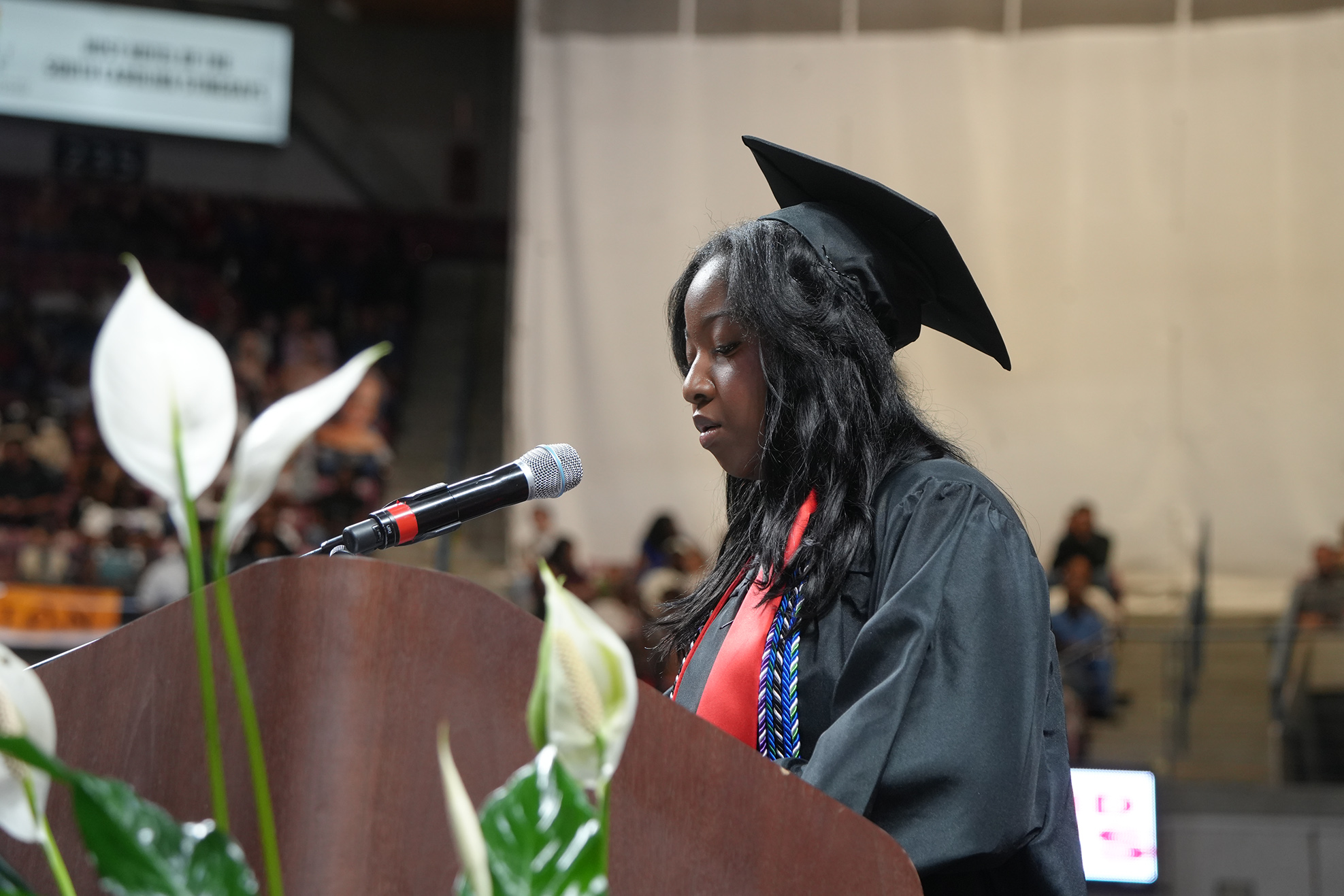 A woman in a black graduation gown and cap speaks at a podium with a microphone.