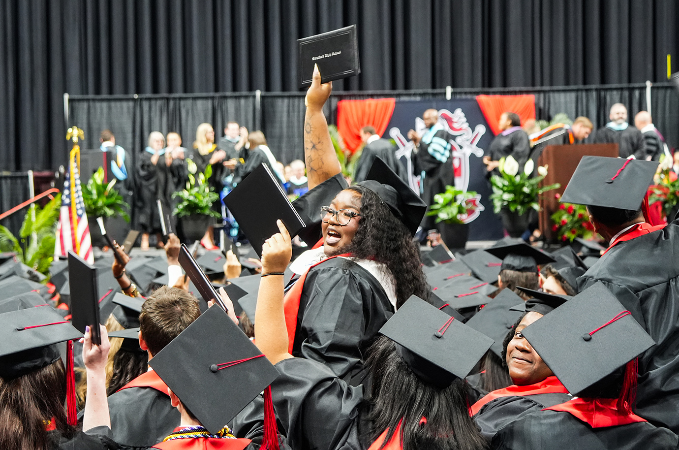 Graduates in black gowns and caps, including one holding a diploma, cheer at an event.