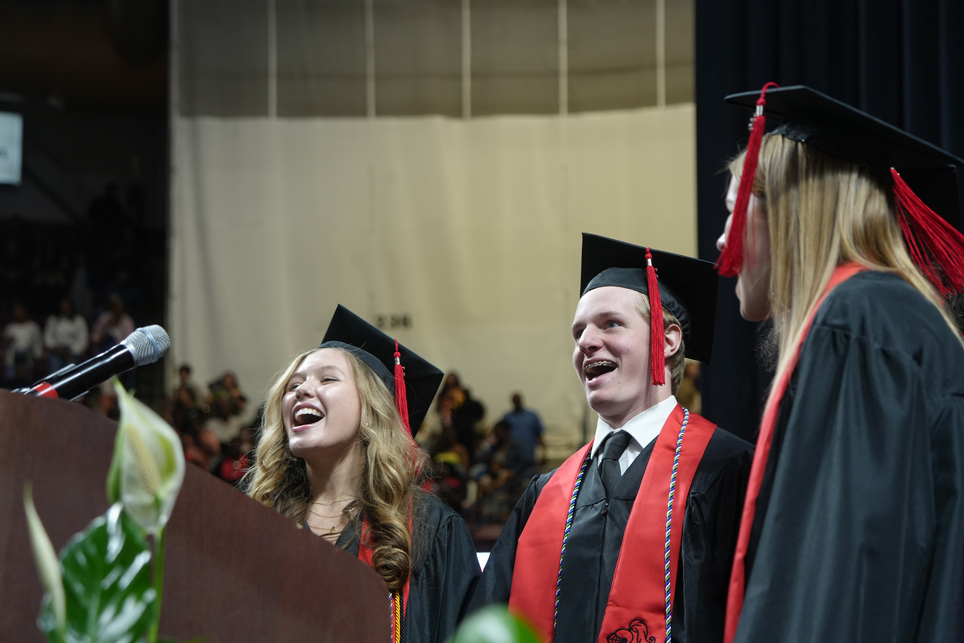 Three graduates in caps and gowns, one laughing, stand near a podium in a large auditorium.