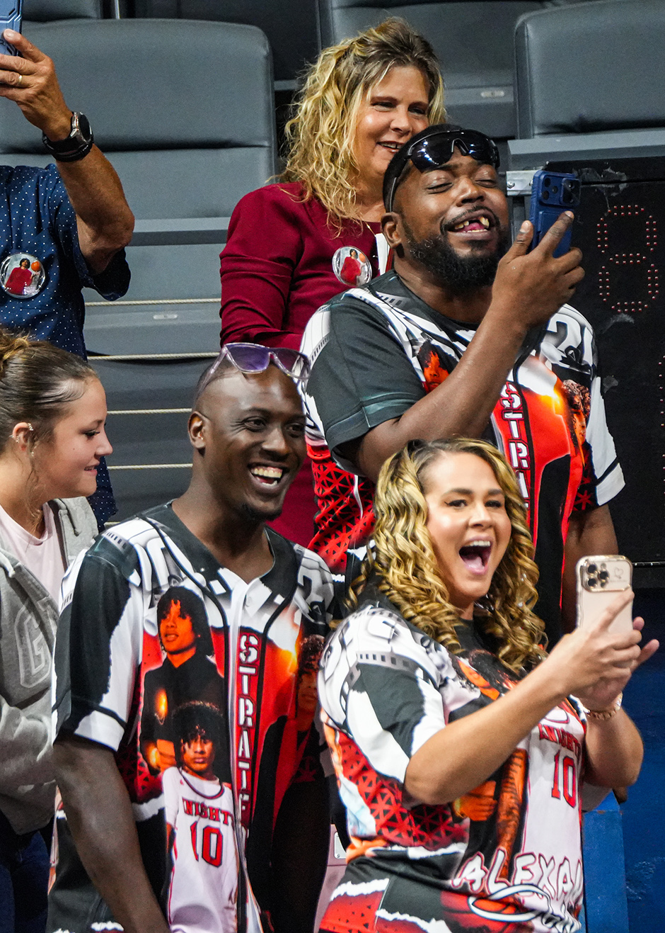 Multiple people standing and smiling in a sports venue. One man wearing a striped shirt holds a cellphone.