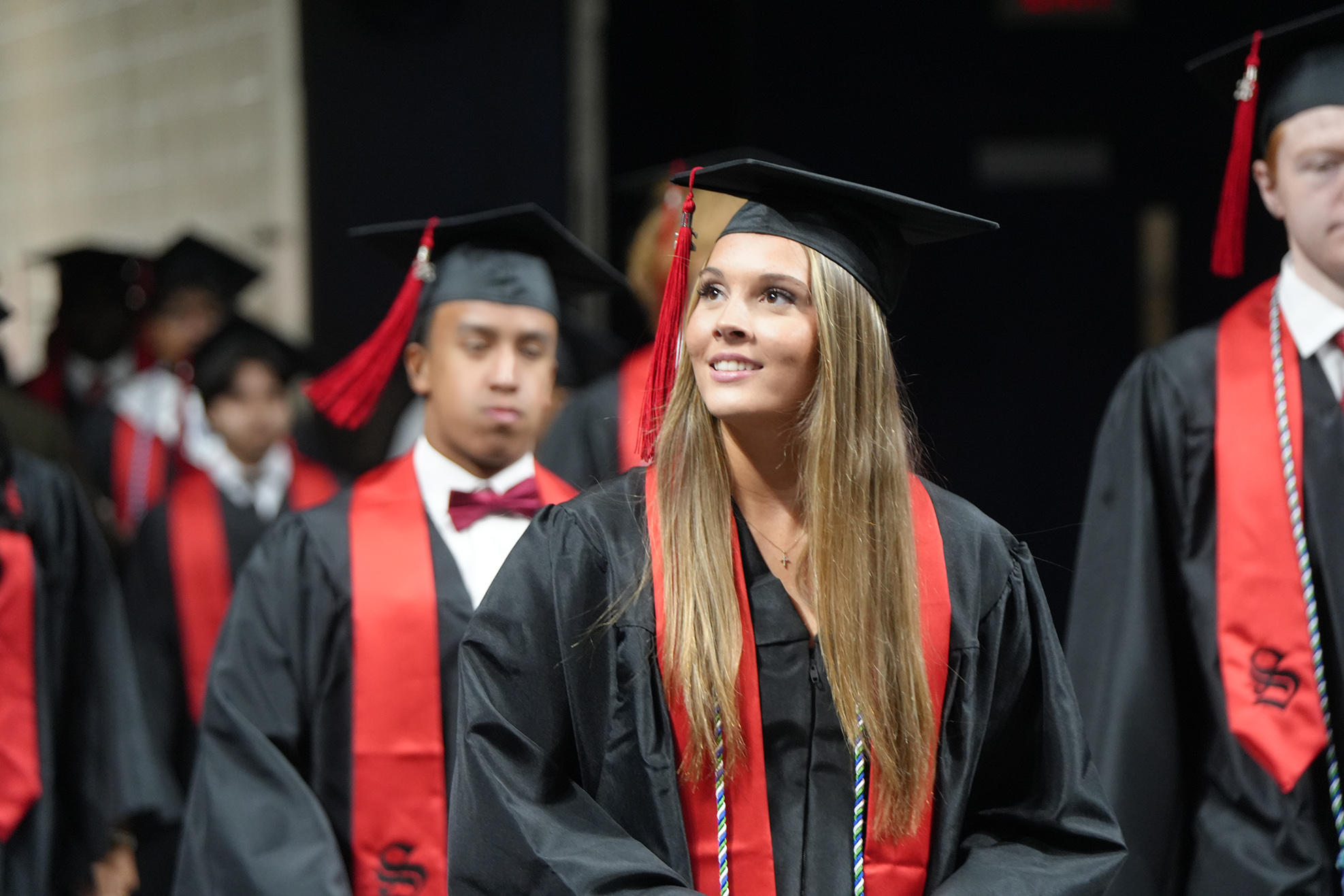 A woman in a black gown with red sash stands in a line of graduates. Some wear red bow ties.