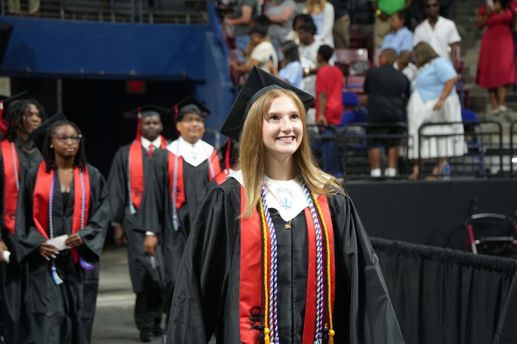 Graduates in caps and gowns walk in a stadium; one smiles at the camera. Behind them, other graduates and spectators in a partially filled bleachers.