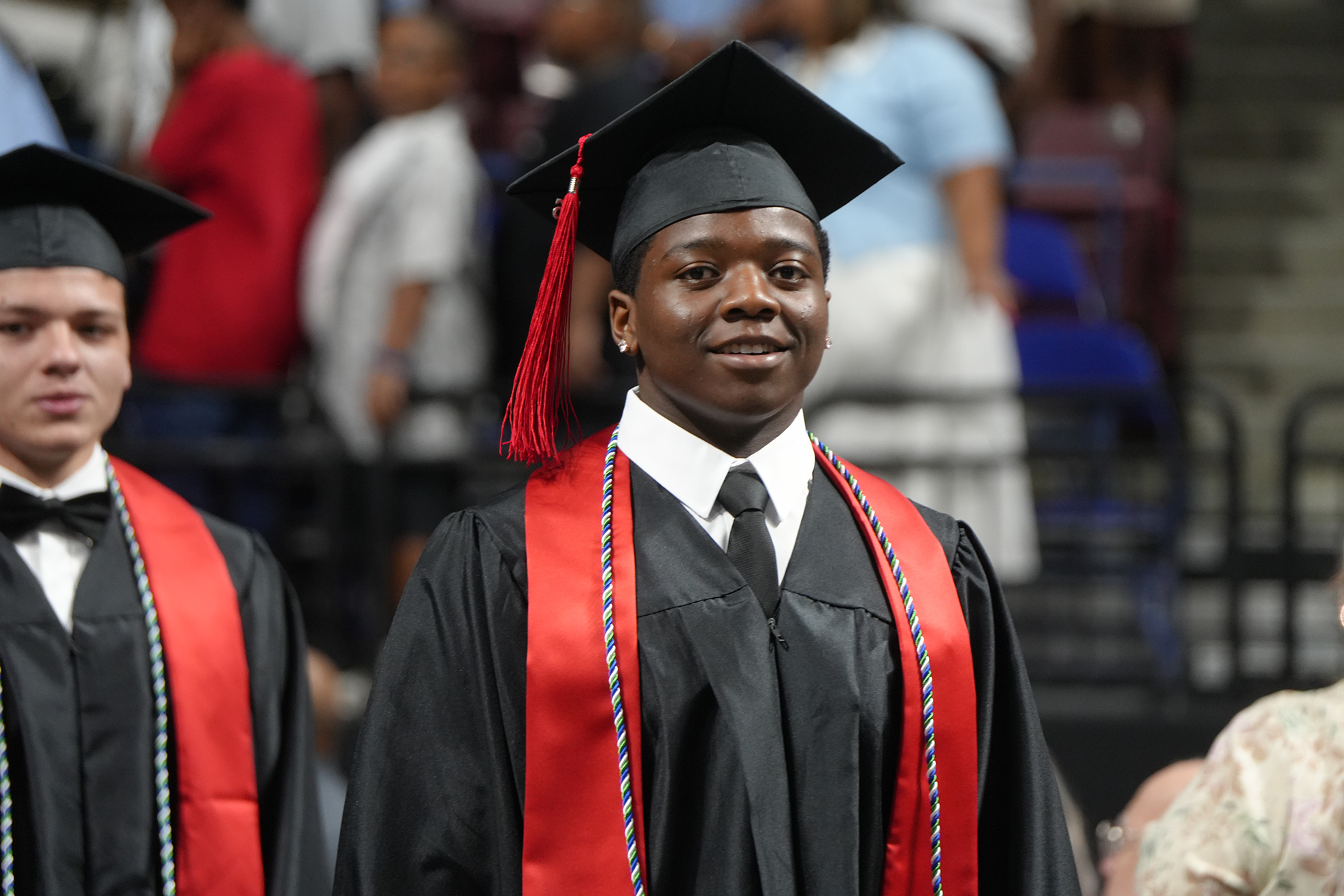 A man in a graduation gown and cap stands, facing the crowd. Another person in a similar outfit is beside him.
