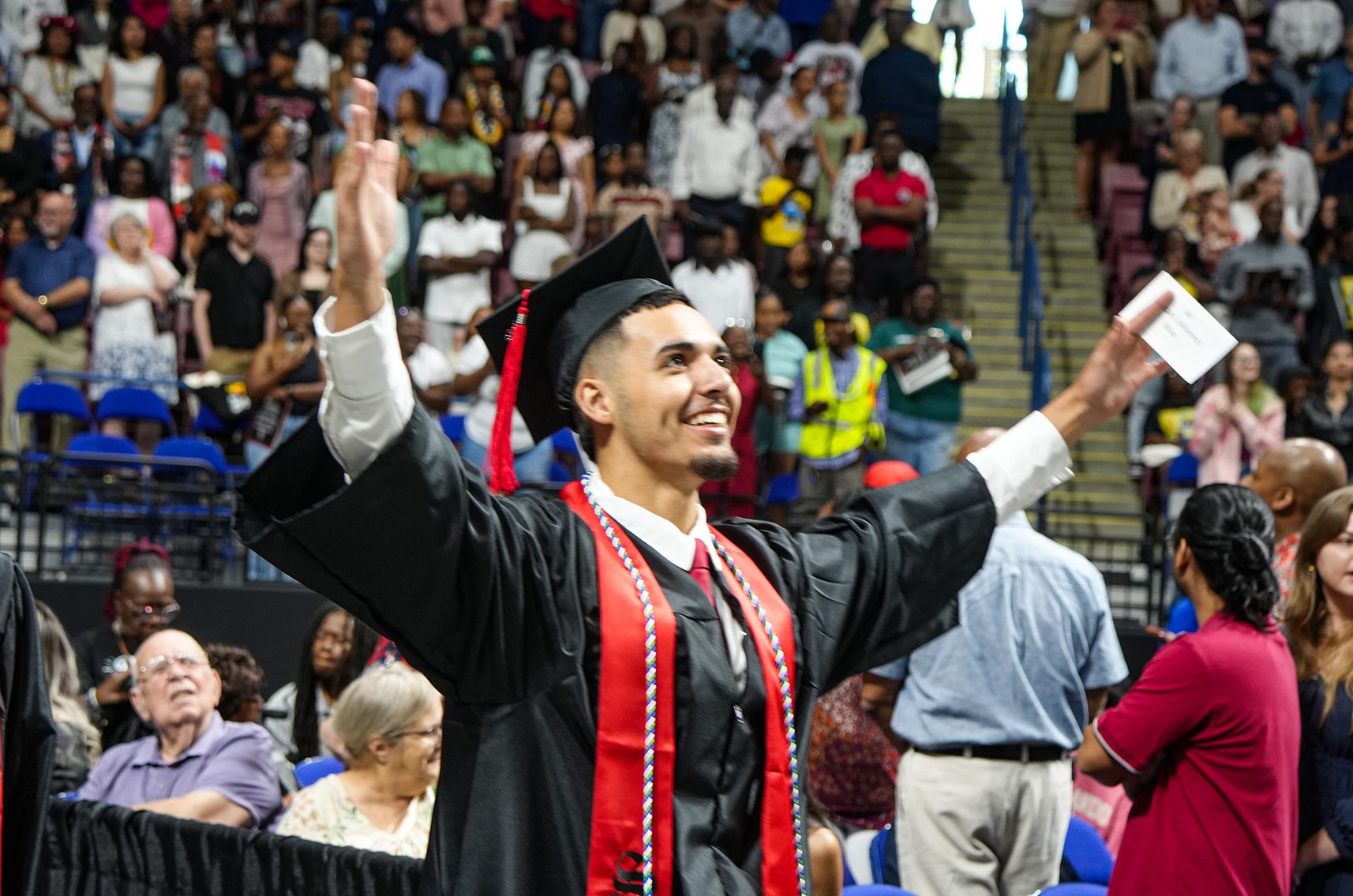 A graduate celebrates in front of a cheering crowd in a stadium. He has his cap in the air.