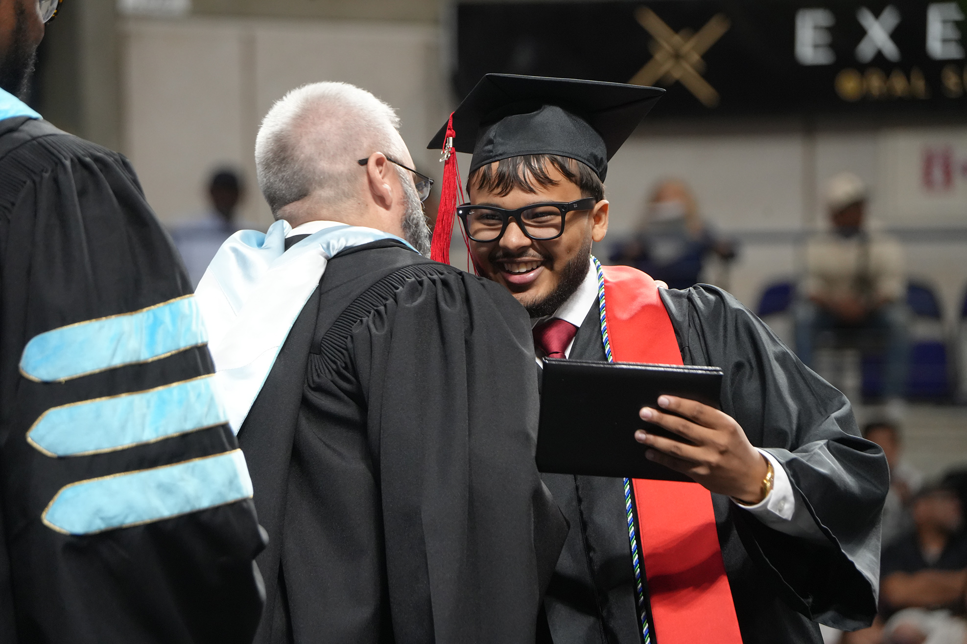 A man in a black graduation gown holds a tablet as another man, also in a gown, looks on.