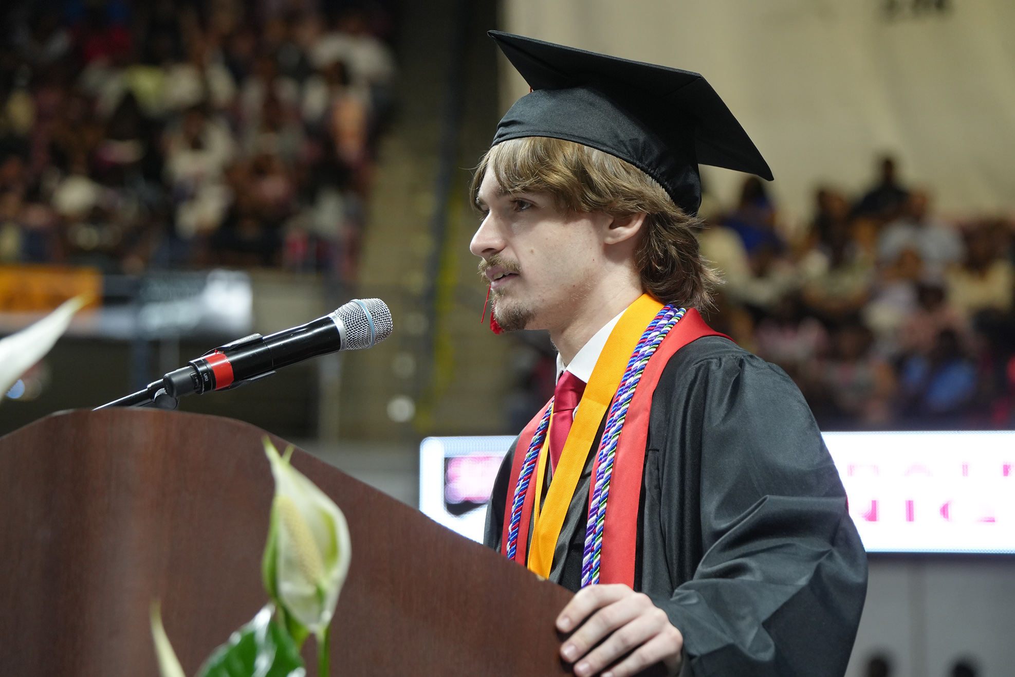 A man with a graduation cap speaks at a podium with a microphone in front of an audience.