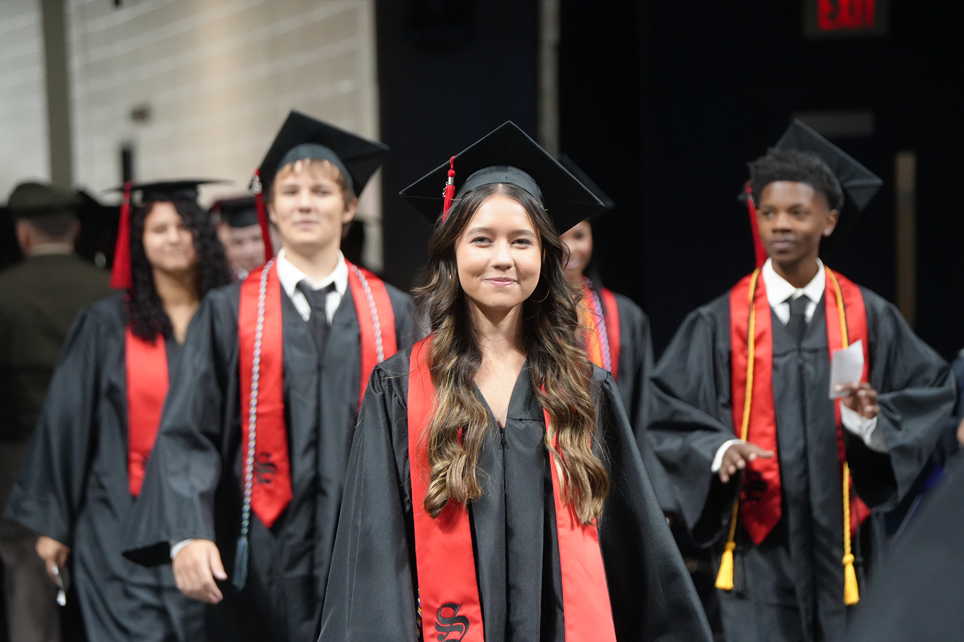 Graduates wearing caps and gowns, with red sashes, walk down a stage at a graduation ceremony.