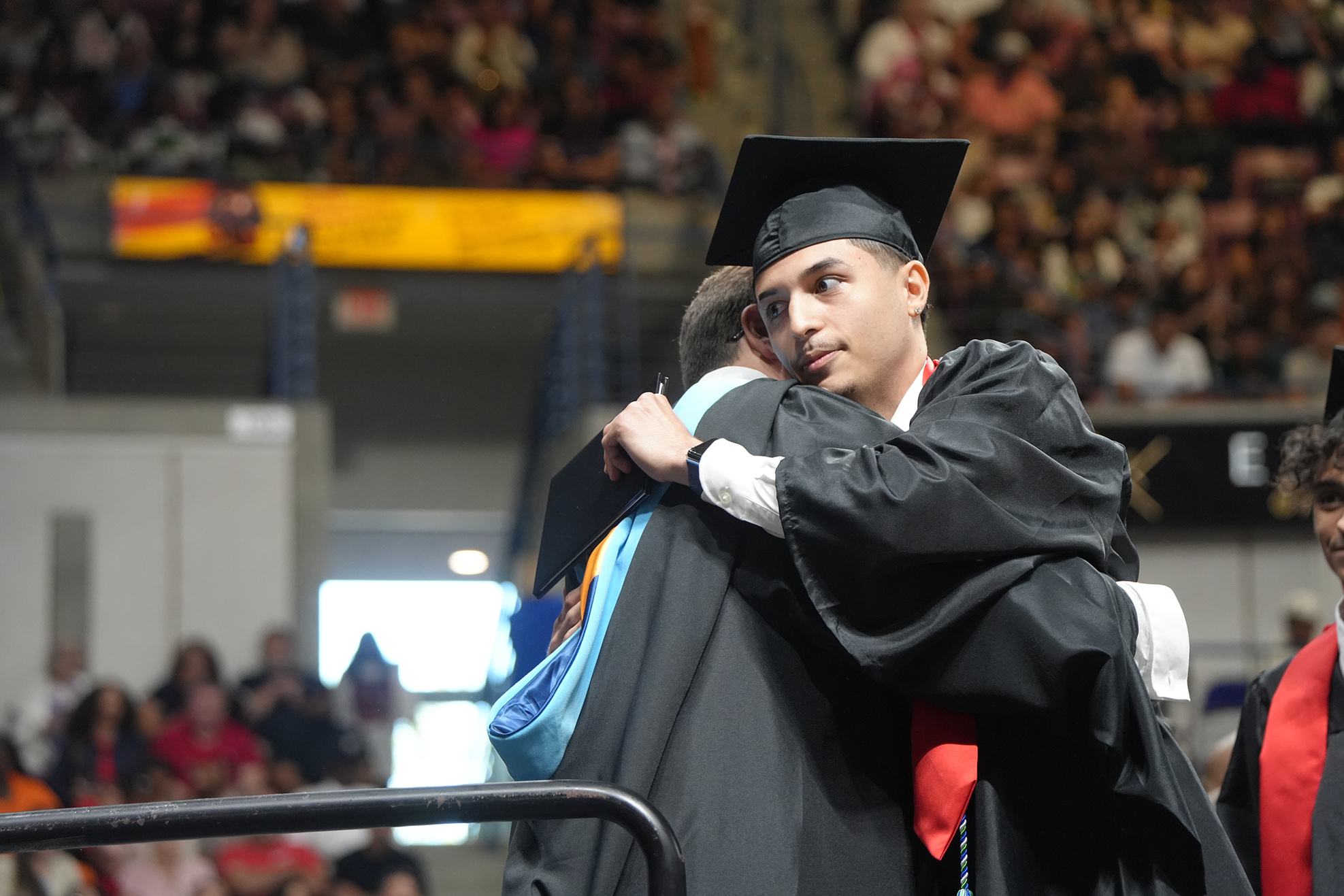 Two people wearing graduation gowns and hats are hugging in front of a crowd in an auditorium.