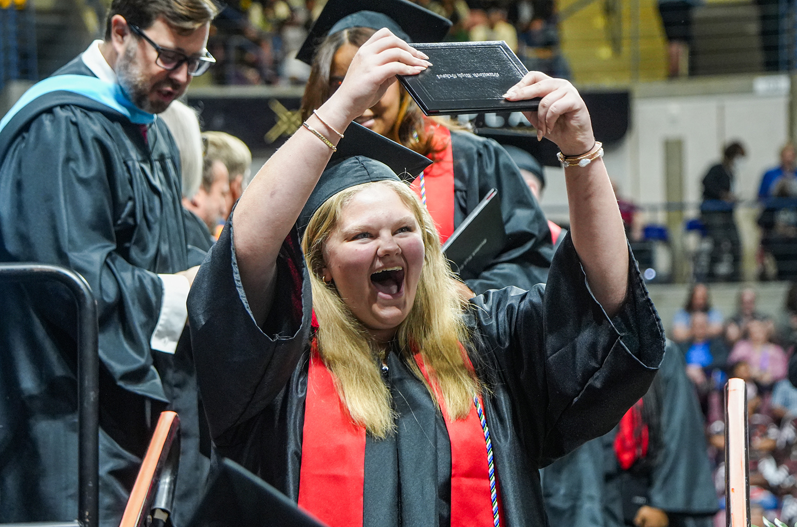 Graduates in gowns at a ceremony. A woman holds a tablet up and smiles. Blurry audience in background.