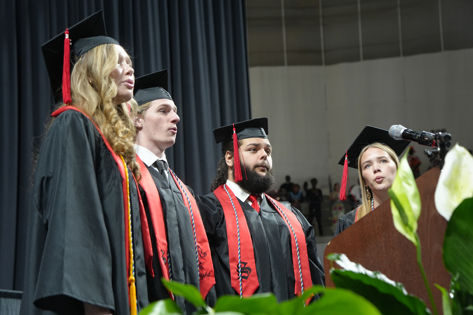 Graduates in academic regalia standing at a podium during a ceremony, with greenery and a microphone in front.