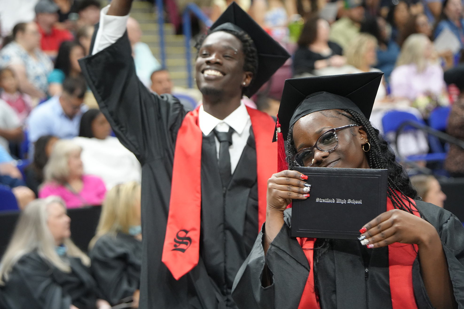 Two graduates in black gowns and caps hold up a folder in front of a crowd.
