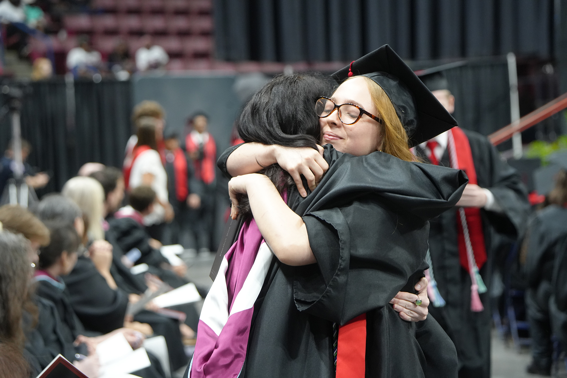 Two graduates in black gowns, one wearing a cap, embrace in an auditorium with others in caps.