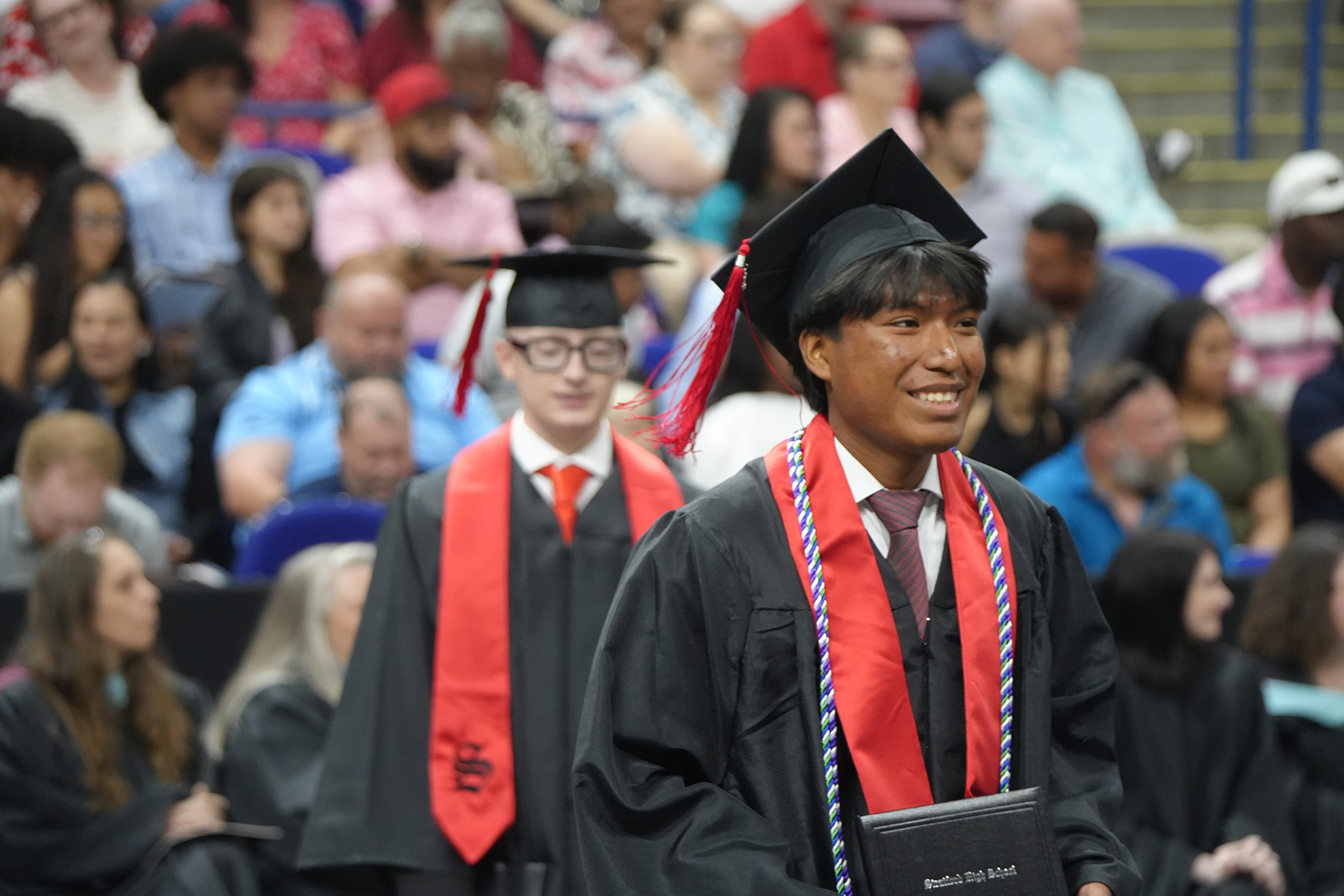 Graduates in academic regalia stand in a line at an outdoor event with a seated audience.