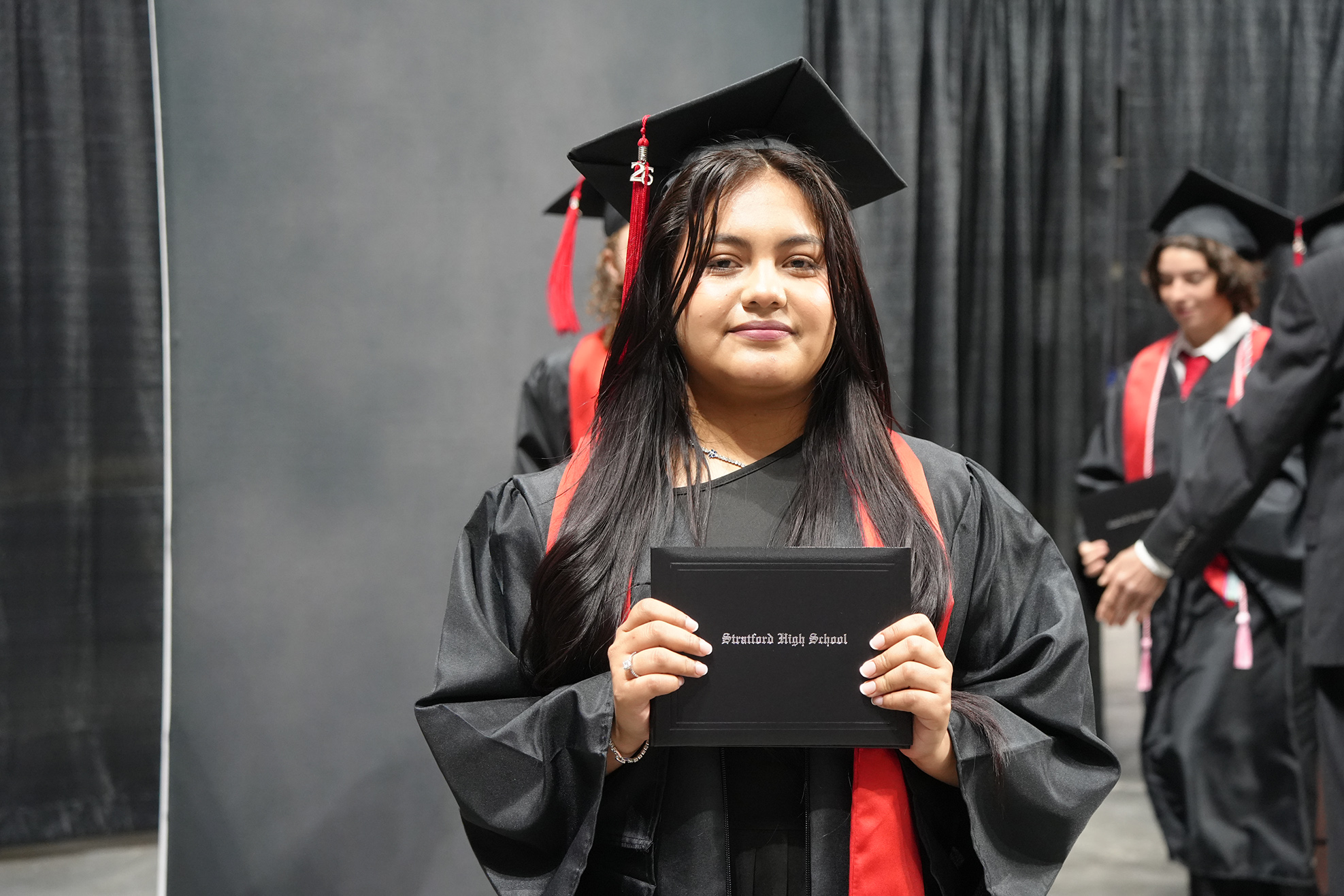 Woman in a black graduation gown holds a diploma while looking forward. Behind her, other graduates in gowns walk.