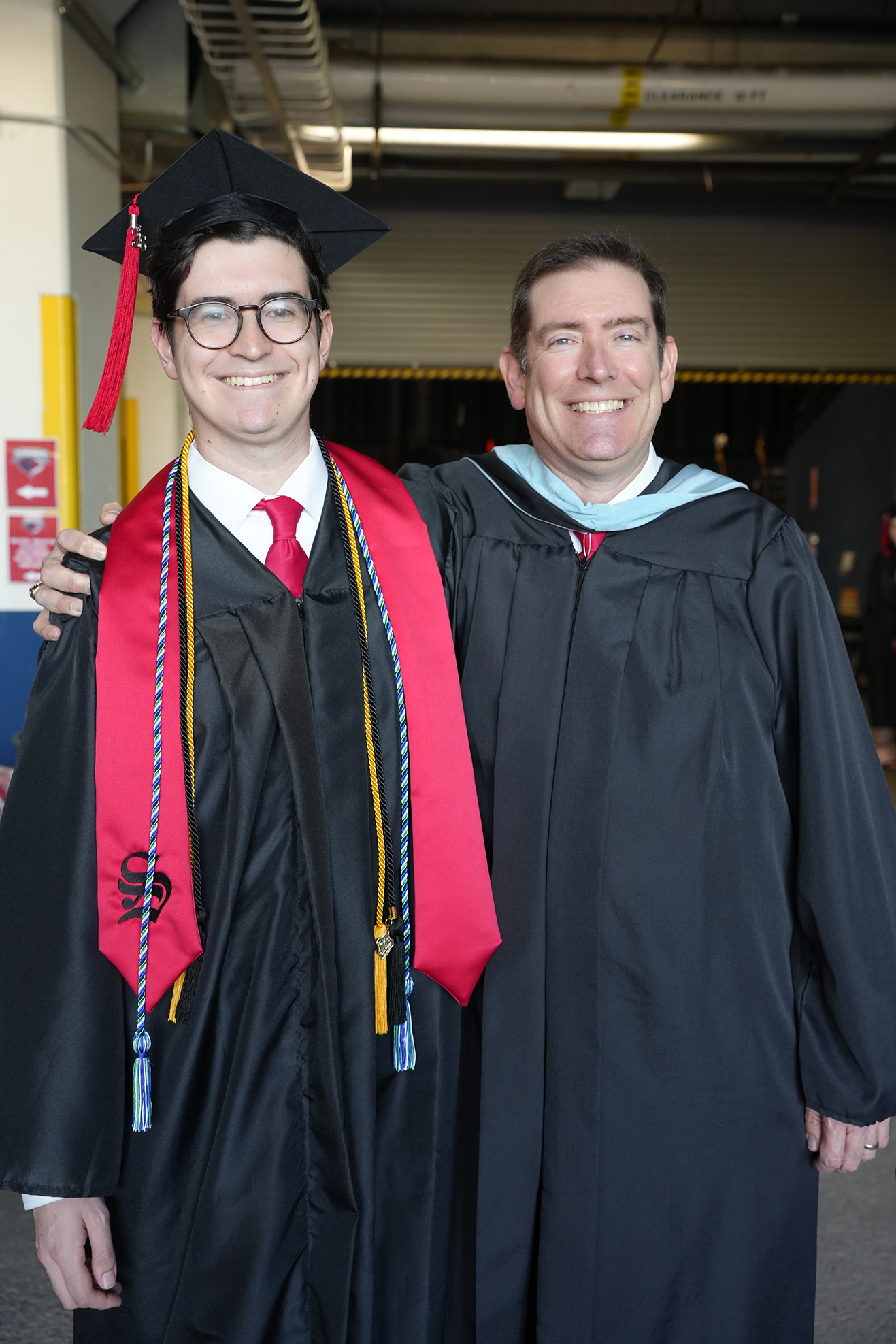 Two men in black graduation gowns and caps, one wearing a red stole, pose for a photo.