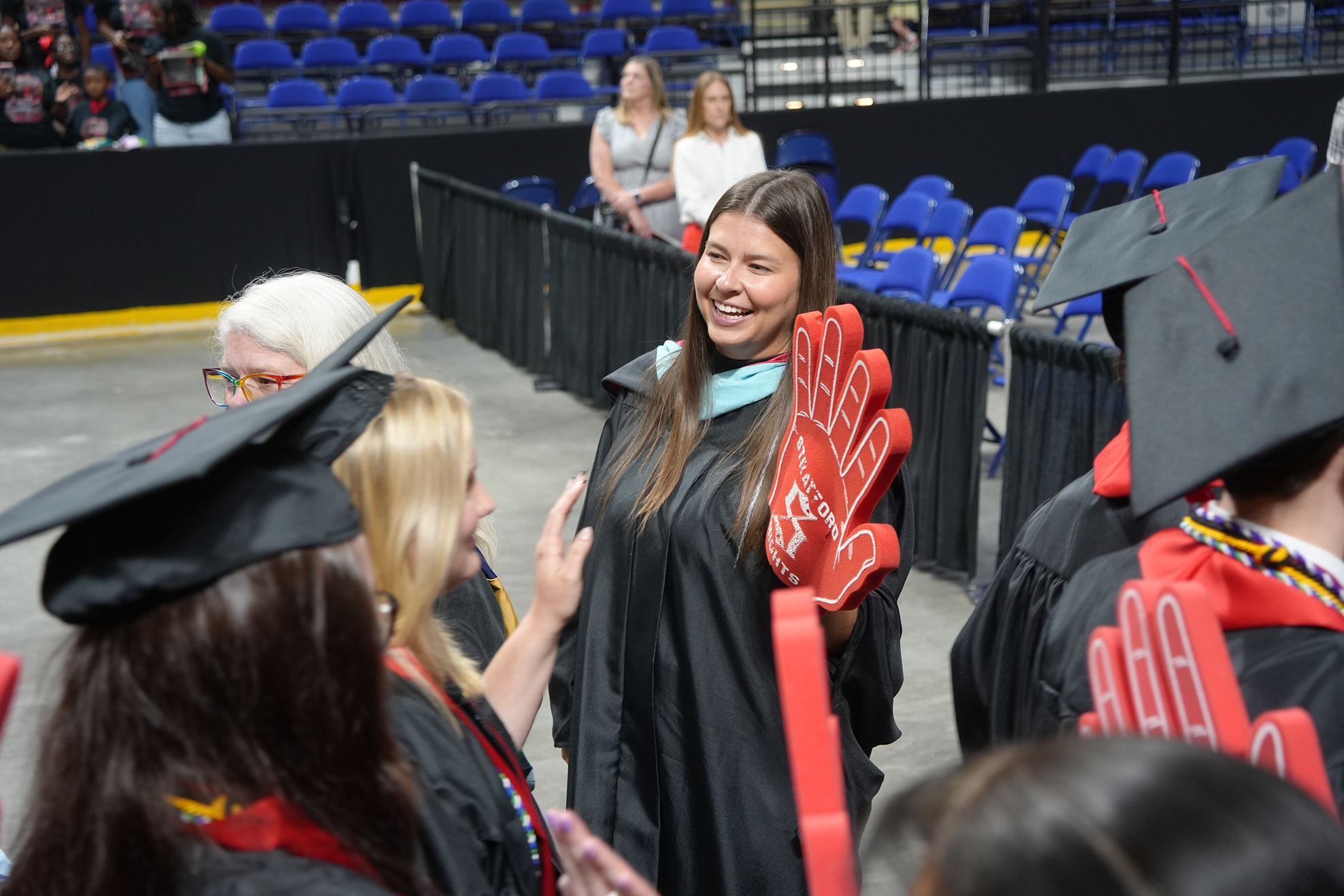 People in graduation attire. A woman smiles, holds red gloves, and speaks to others. Behind, rows of blue chairs.