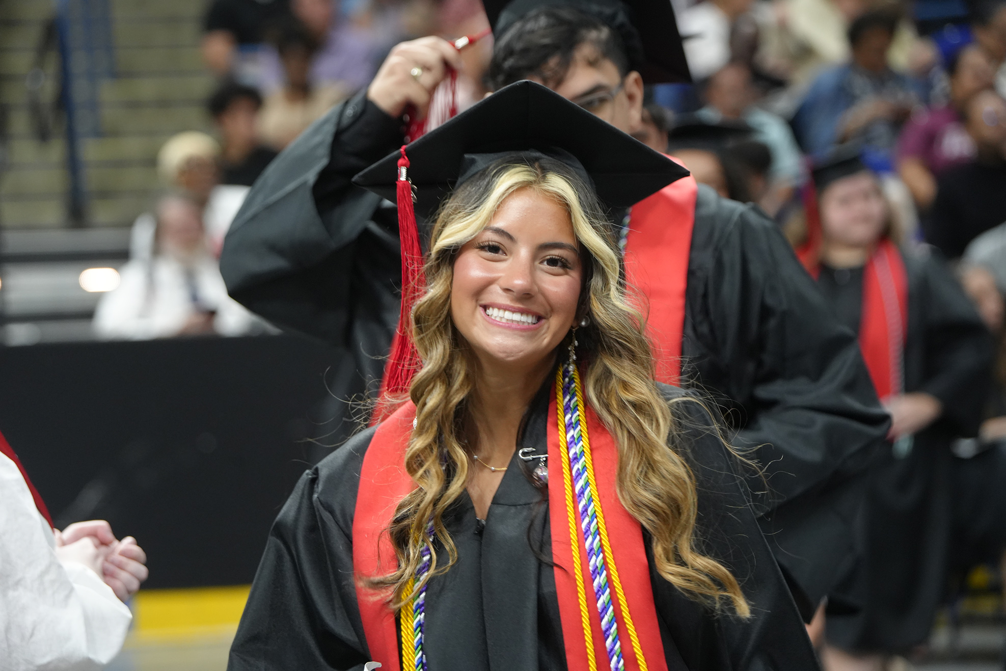 Woman in black graduation gown with red and blue stole, smiling, walking in front of audience.