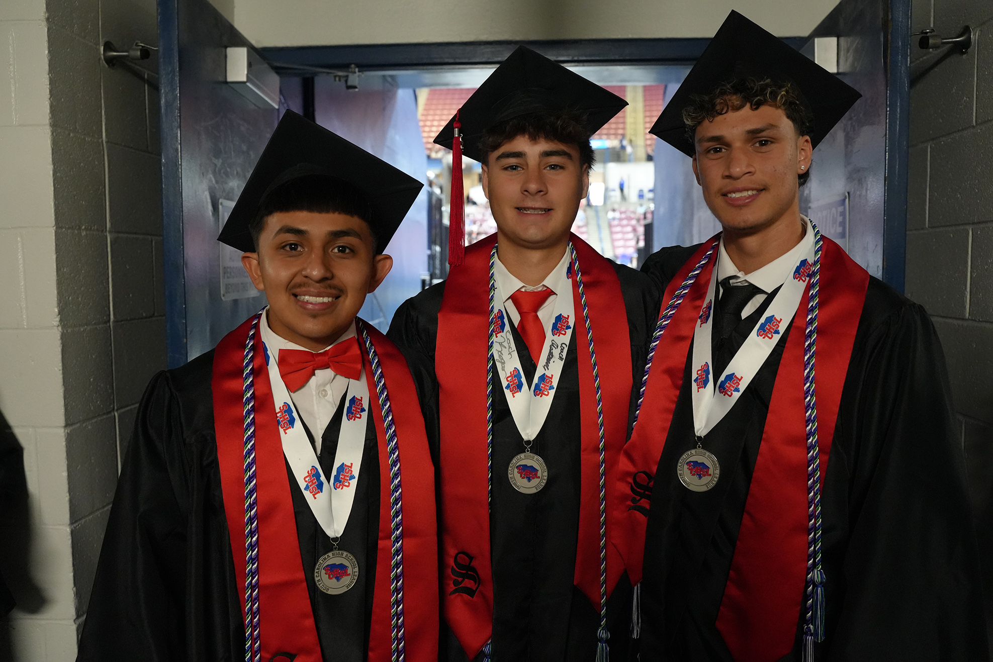 Three young men in graduation robes with red sashes, black caps, and medals stand in a doorway.