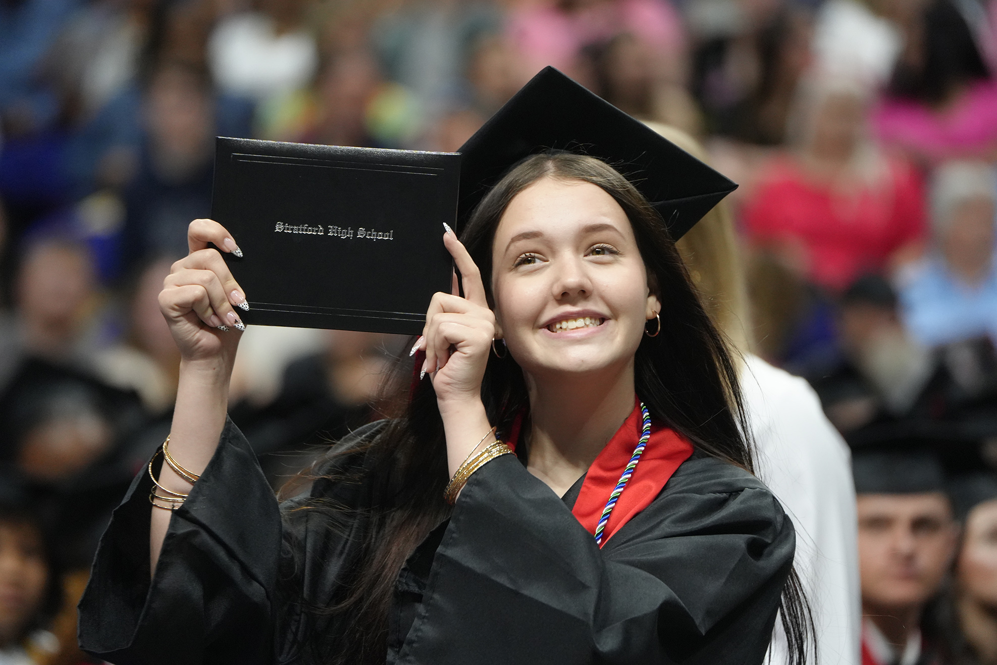 Woman in a black graduation gown holds her diploma and smiles at the camera during a ceremony.