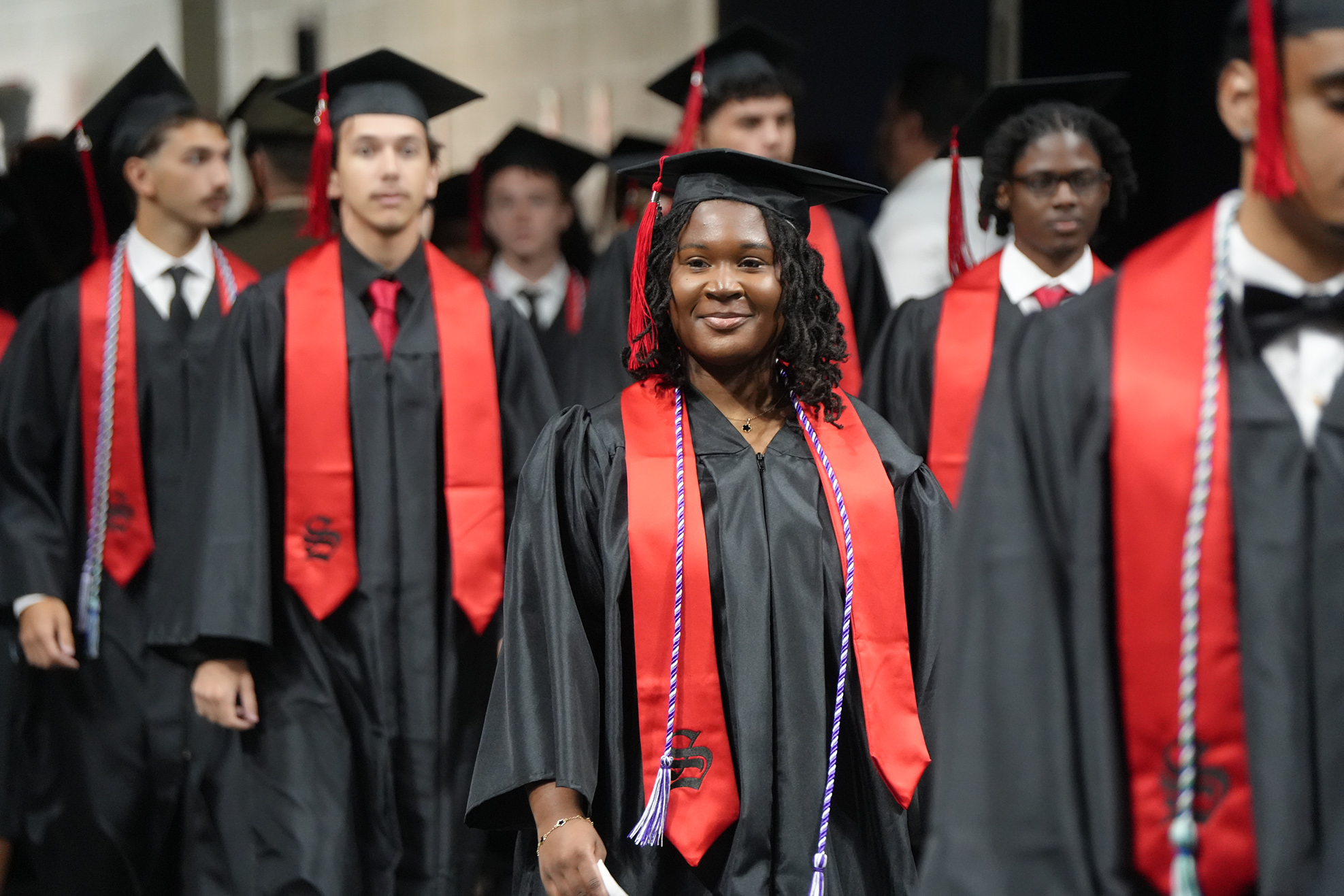Graduates in black gowns with red sashes walk down a stage, some with caps. A woman smiles.