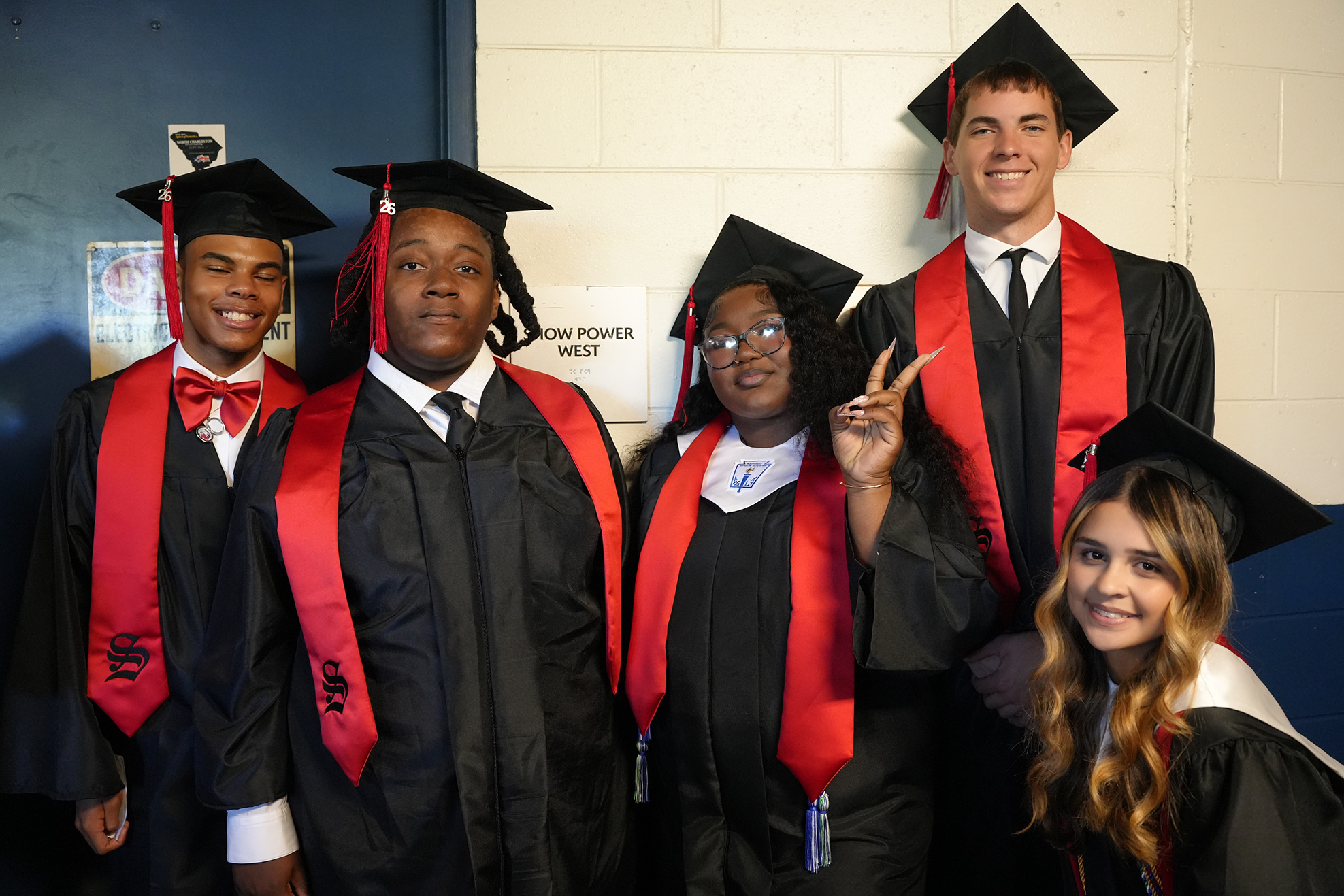 Five people in graduation gowns pose for a photo. Three wear red stoles, two with black caps. The background has a white wall and a sign.
