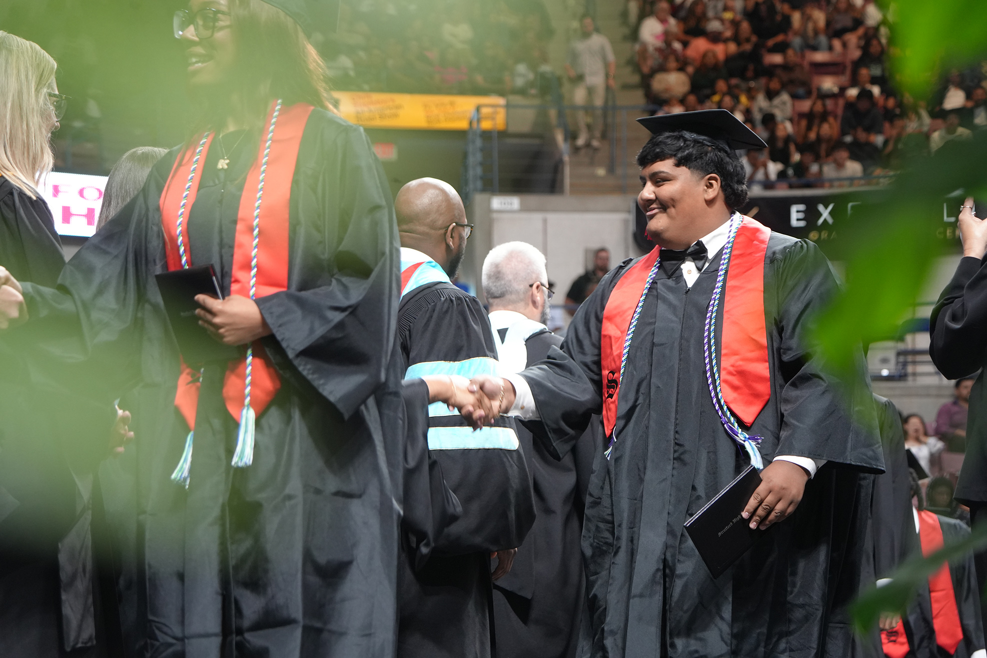 Graduates in black caps and gowns shake hands with each other in a stadium. Some people are seated in the background.