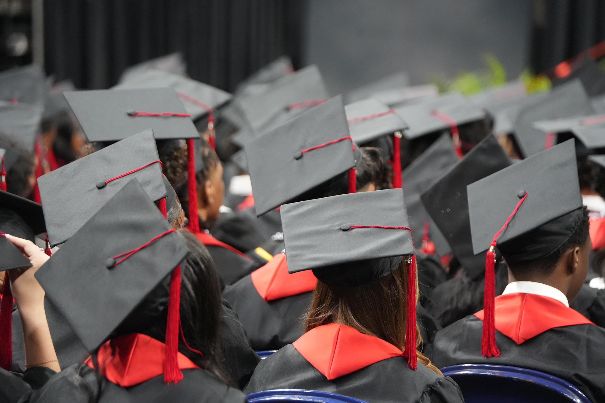 People in black graduation gowns and caps with red tassels sit in rows at a ceremony.