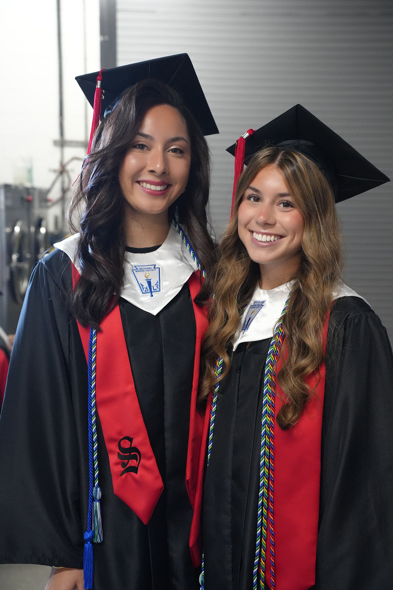 Two women in black and red graduation gowns and hats stand close, smiling, against a gray background.