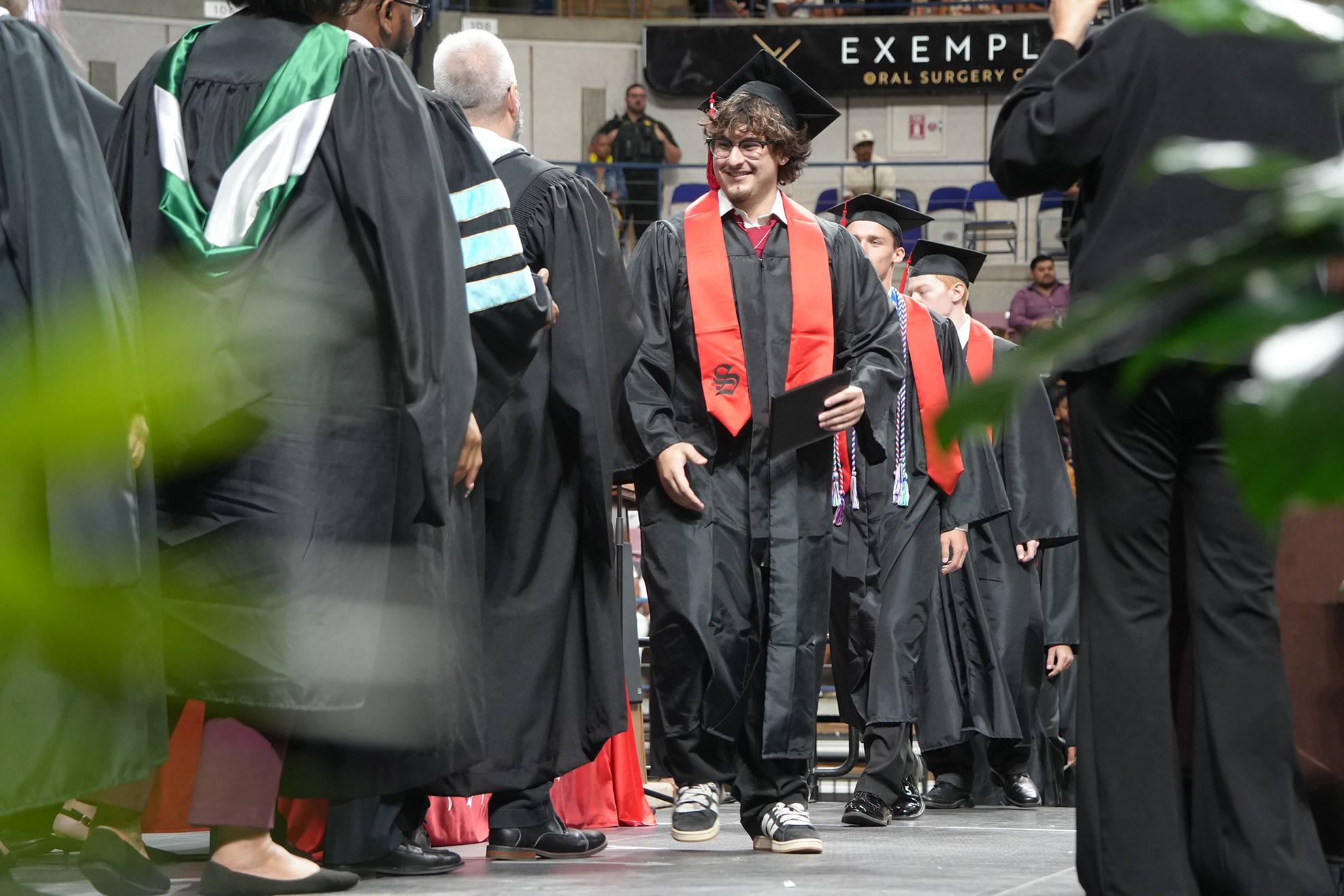 Several people in academic gowns and hats walk across a stage. One person holds a diploma.