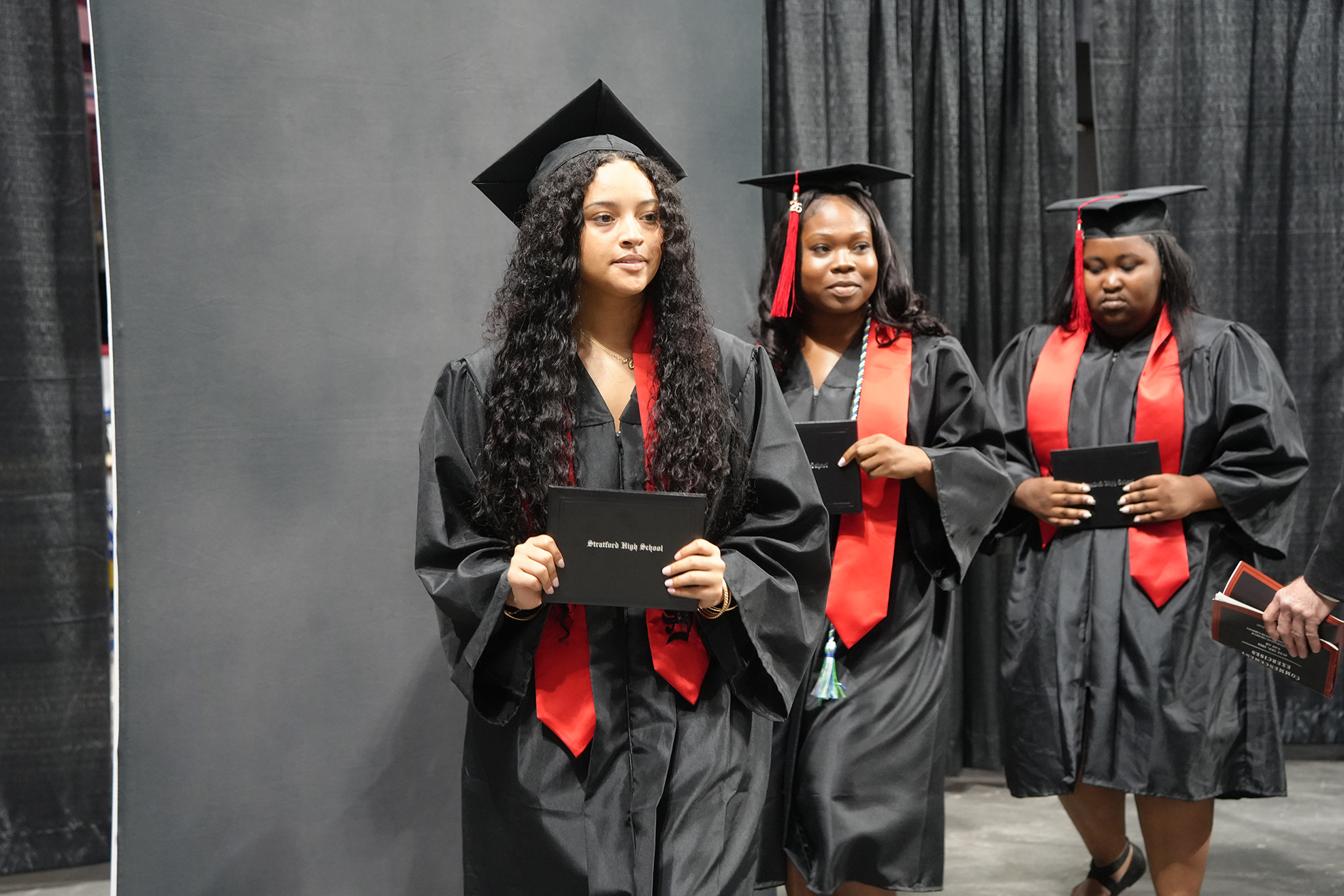 Three women in black graduation gowns and caps, each holding a diploma, stand side by side.