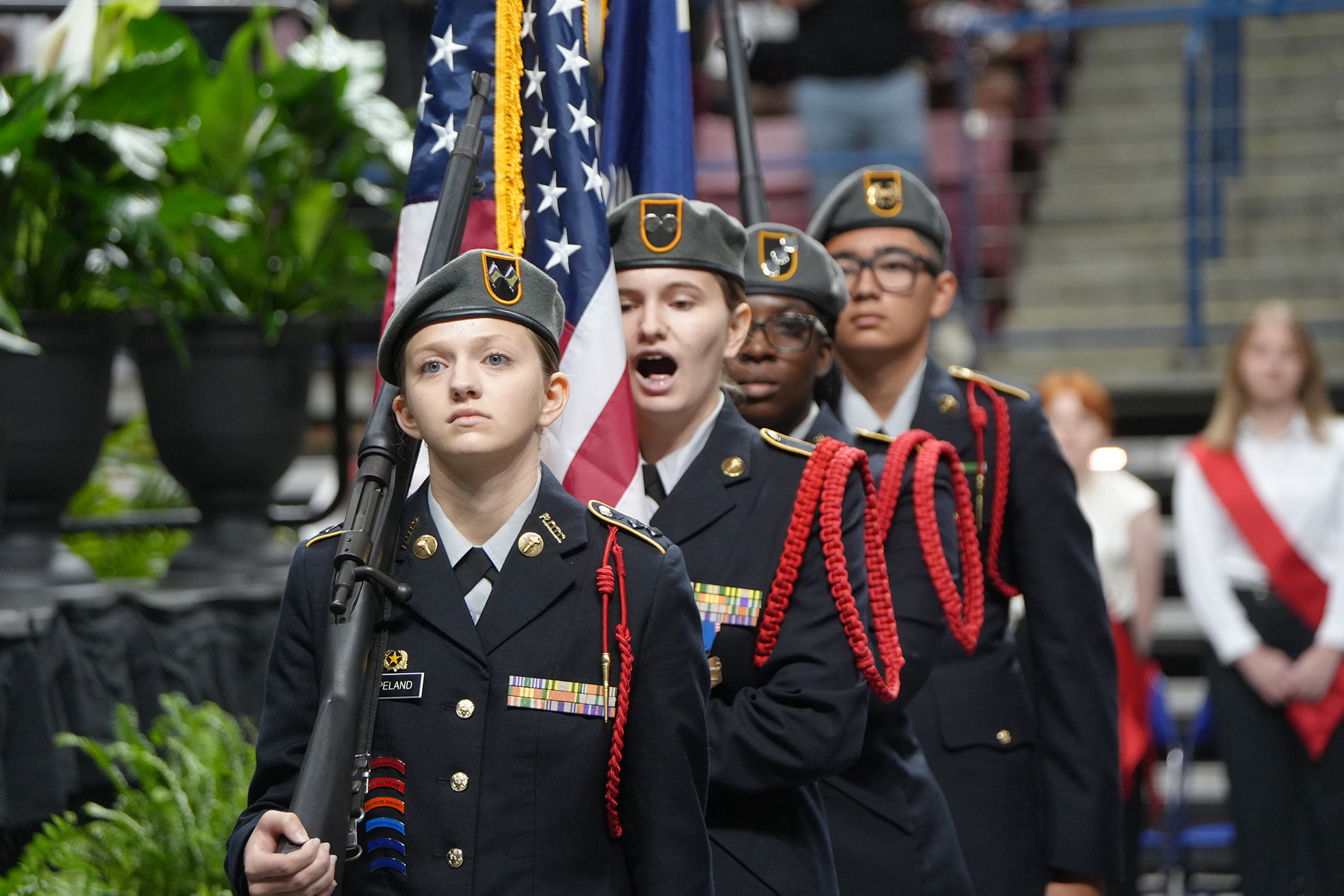 Five uniformed individuals, holding an American flag and a rifle, march in a row. They wear hats and glasses.