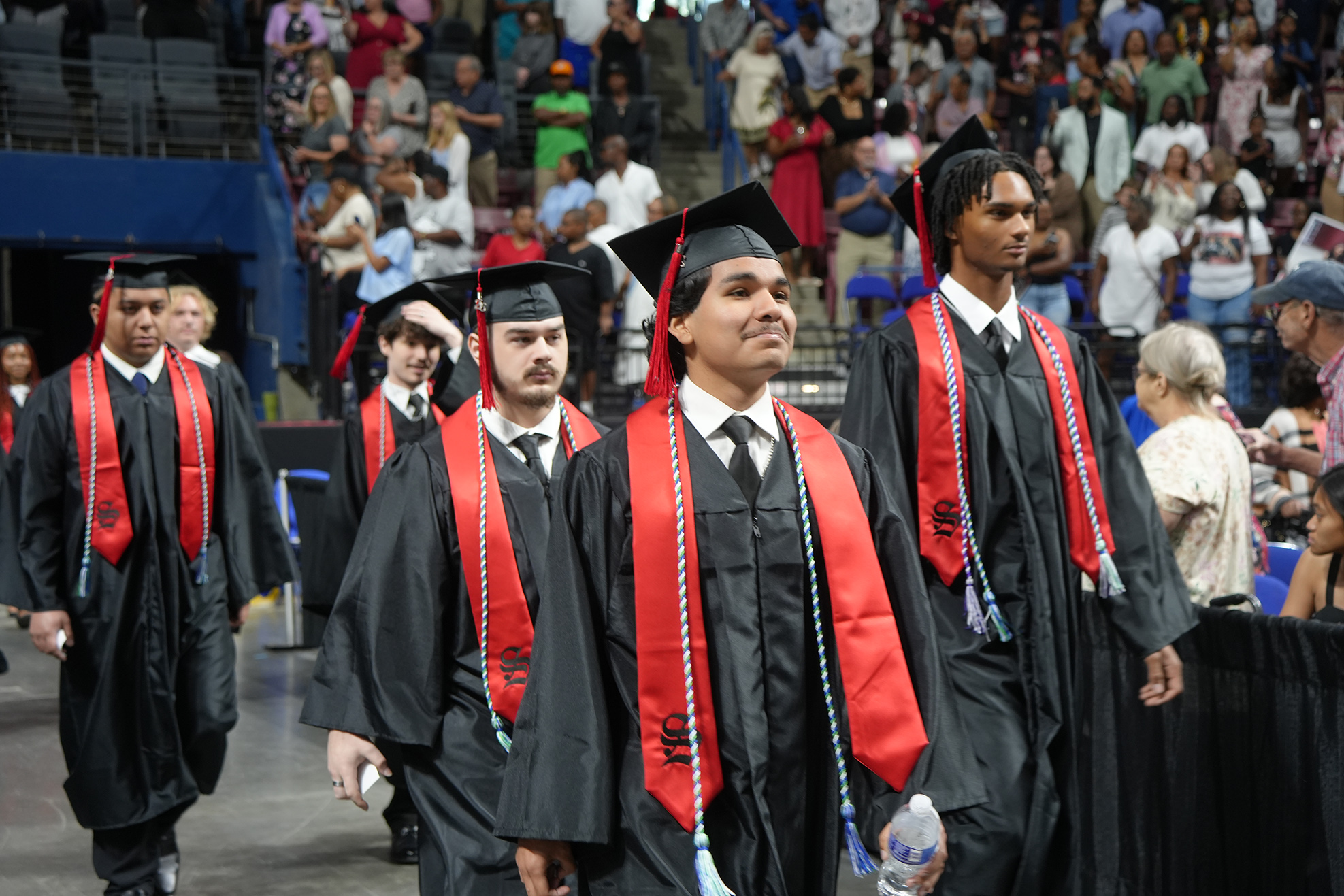 Graduates in black gowns and caps walk in a ceremony. Spectators watch from the stands.